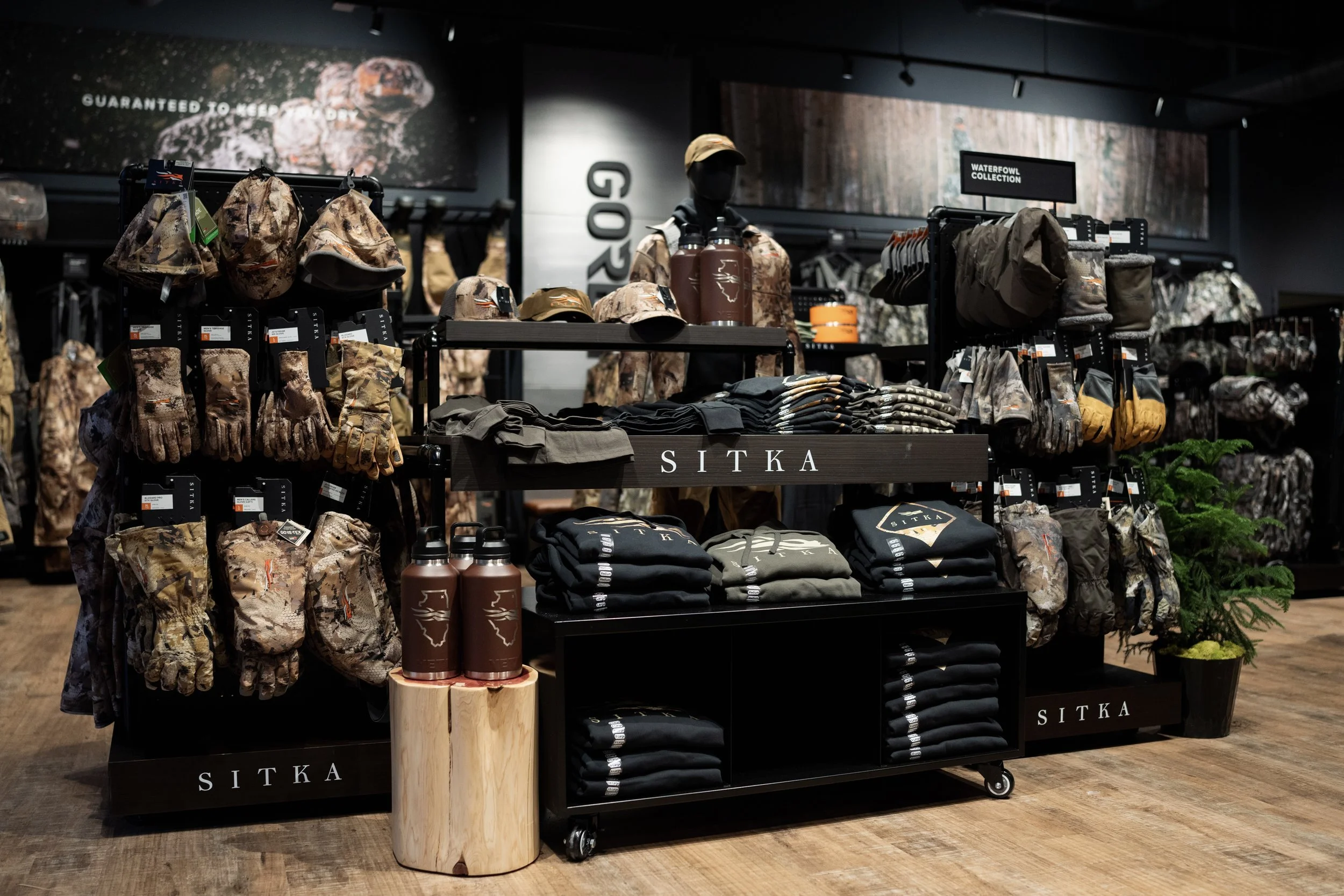 Display of outdoor clothing and gear, including camouflage hats, gloves, jackets, and water bottles, in a retail store section labeled 'Waterfowl Collection' and 'SITKA' branding.