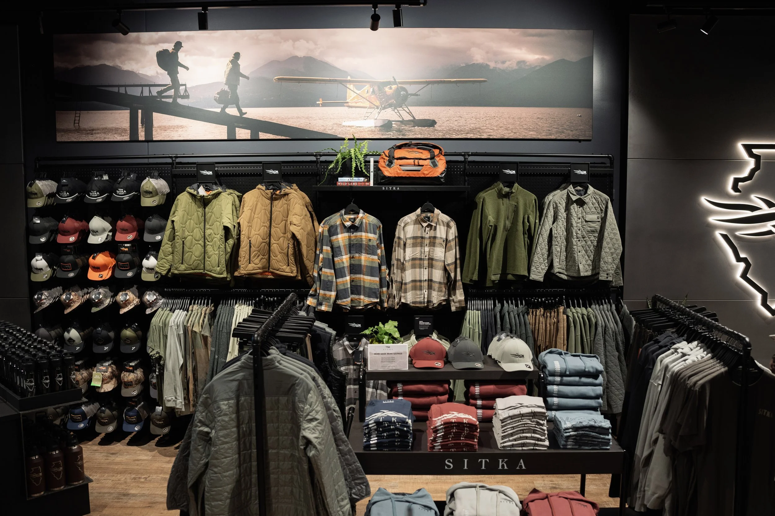 Store display featuring outdoor apparel such as jackets, hats, and shirts, with a large nature-themed photograph on the wall showing hikers and a seaplane on water with mountains in the background.