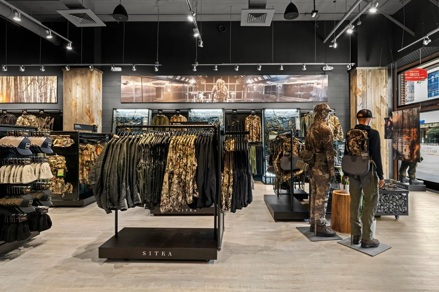 Military apparel and accessories displayed in a retail store, with mannequins dressed in camouflage outfits, racks of jackets, hats, and a wall of digital screens in the background.