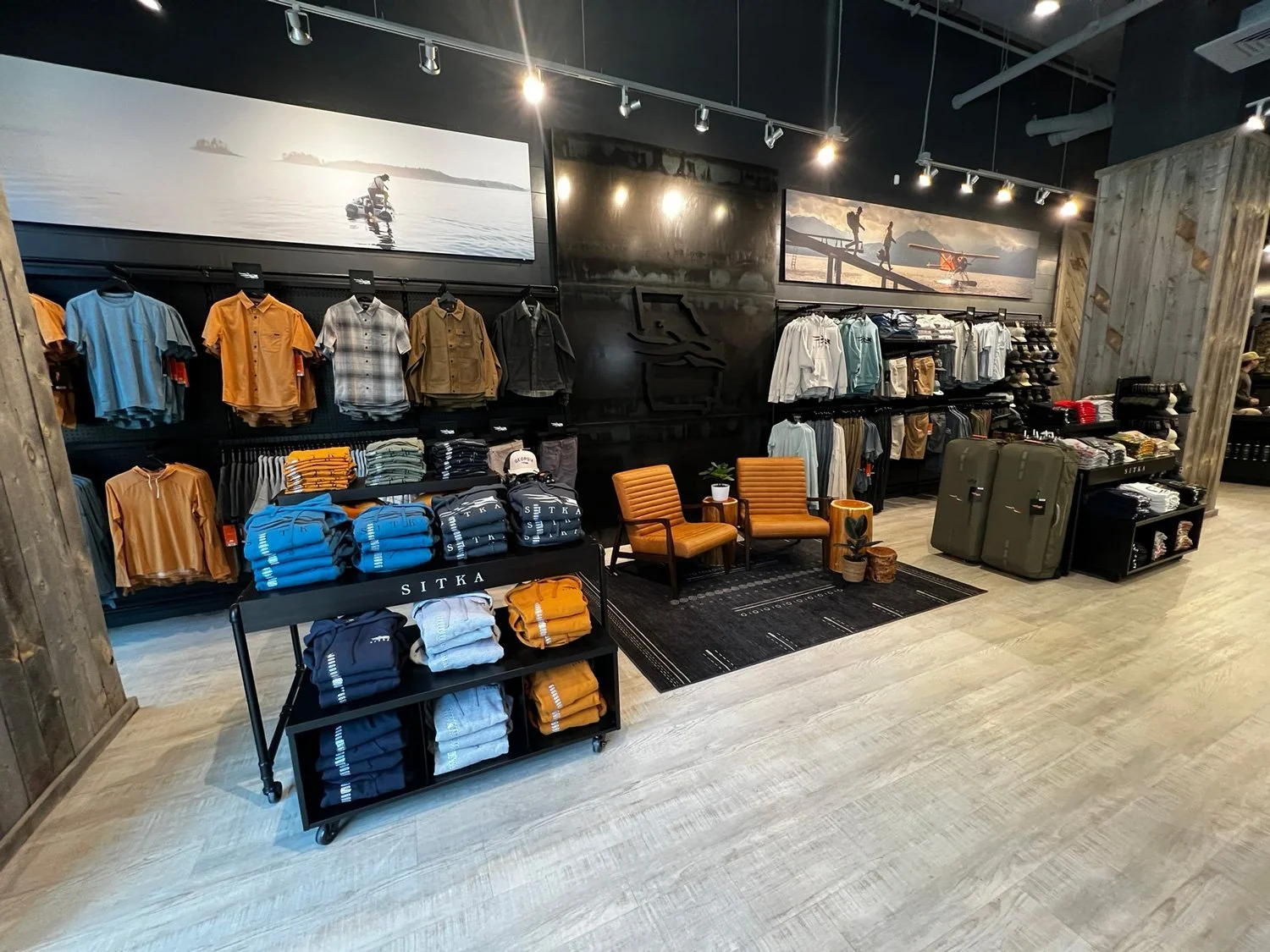 Clothing store display with casual apparel, chairs, luggage, and wall art of outdoor scenes.
