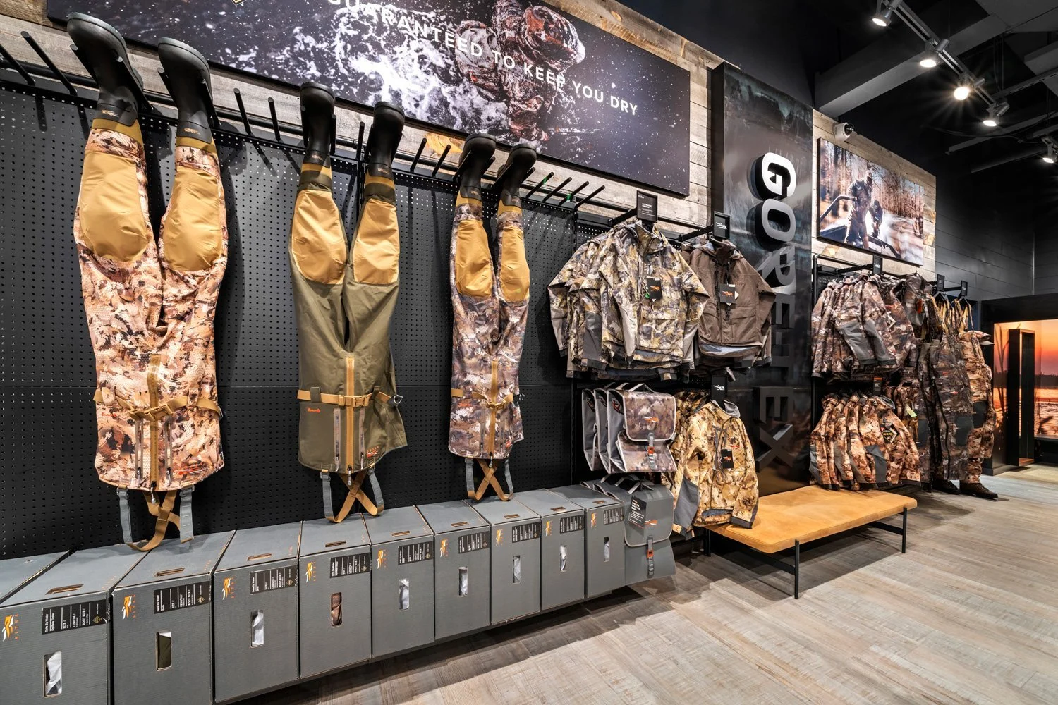 Store display of camouflage outdoor gear, including jackets and pants, hanging on a black pegboard wall with boxes below and large outdoor-themed posters on the wall.