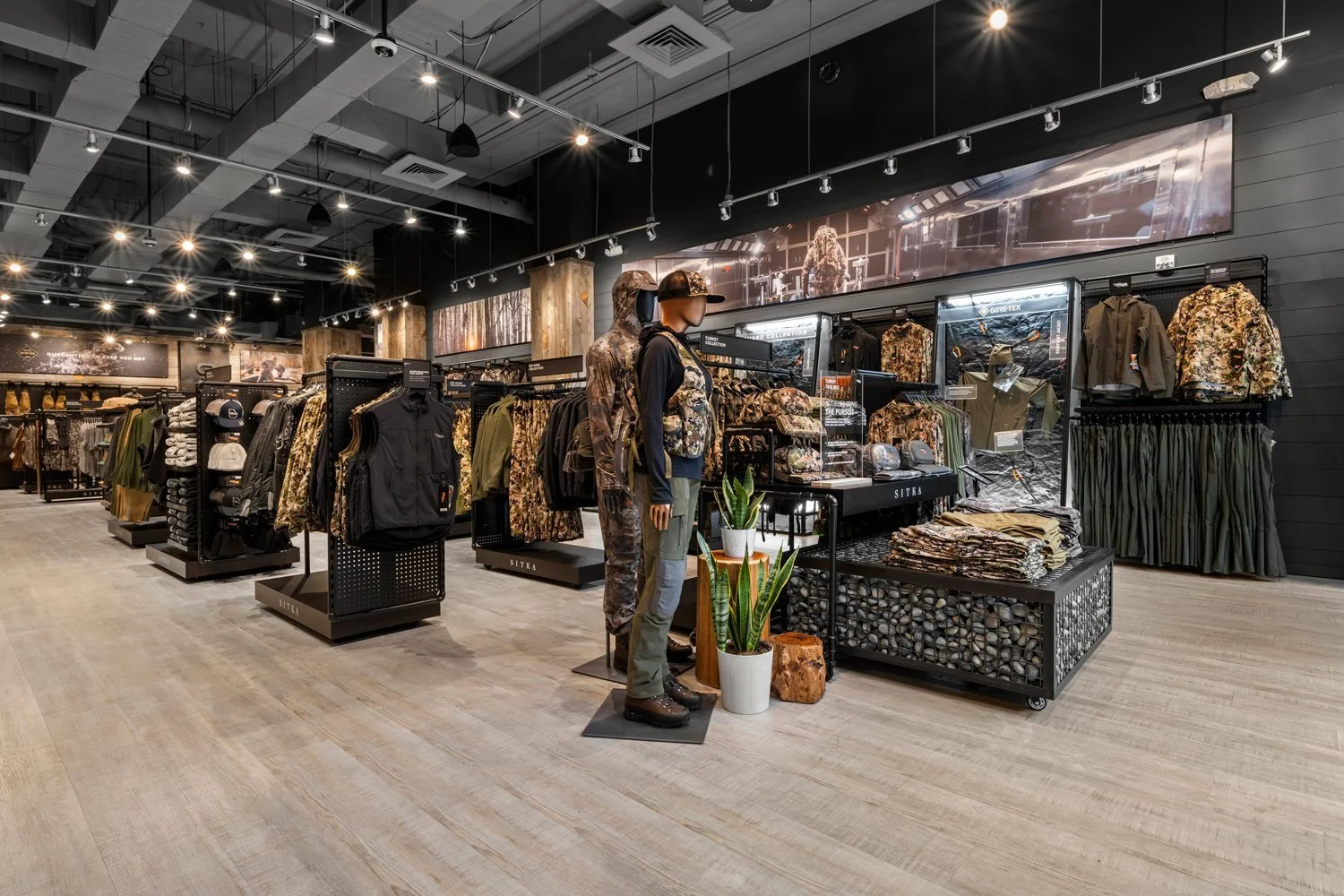 Interior of a retail store selling outdoor and camouflage clothing and gear, with mannequins dressed in outdoor apparel and displays of hats, jackets, and pants.