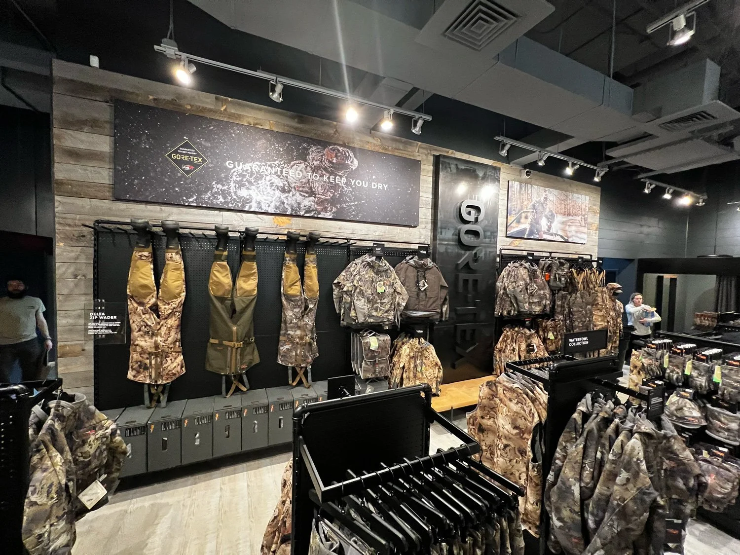 Outdoor clothing store with camouflage jackets, vests, backpacks, and accessories displayed on black and wooden fixtures. Signs indicating waterproof and waterproof collection. Large GORE-TEX sign and promotional banners on wooden wall.