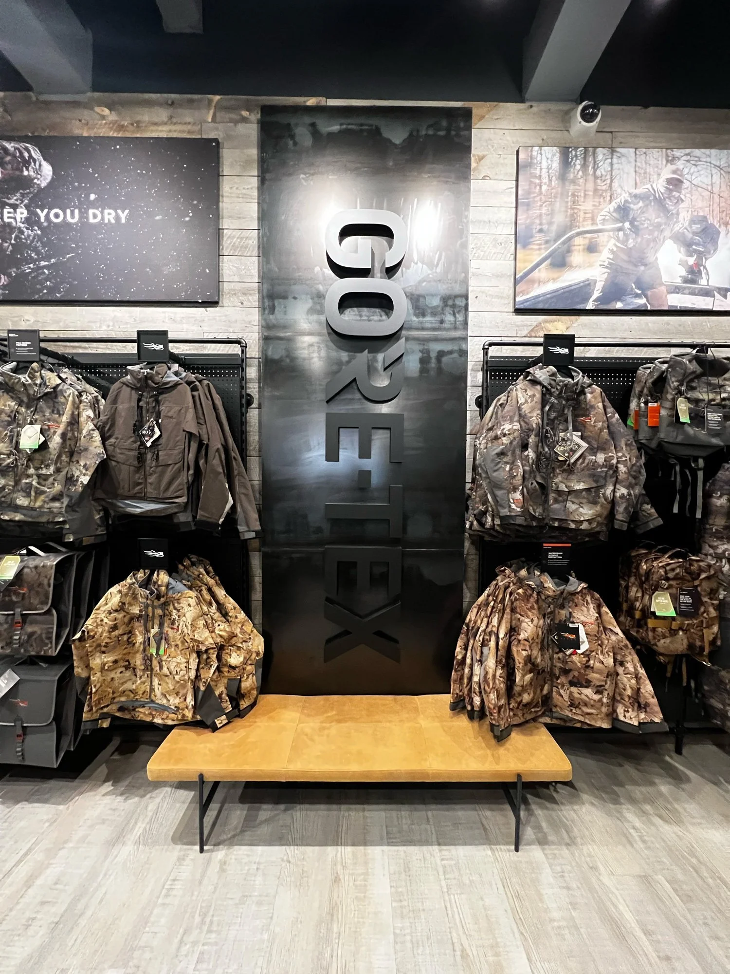 Display of camouflage jackets and backpacks on black racks in a store. Two advertisements are on the wall behind: one with a person wearing camouflage in a winter forest, and another with camouflage gear and a slogan about staying dry.