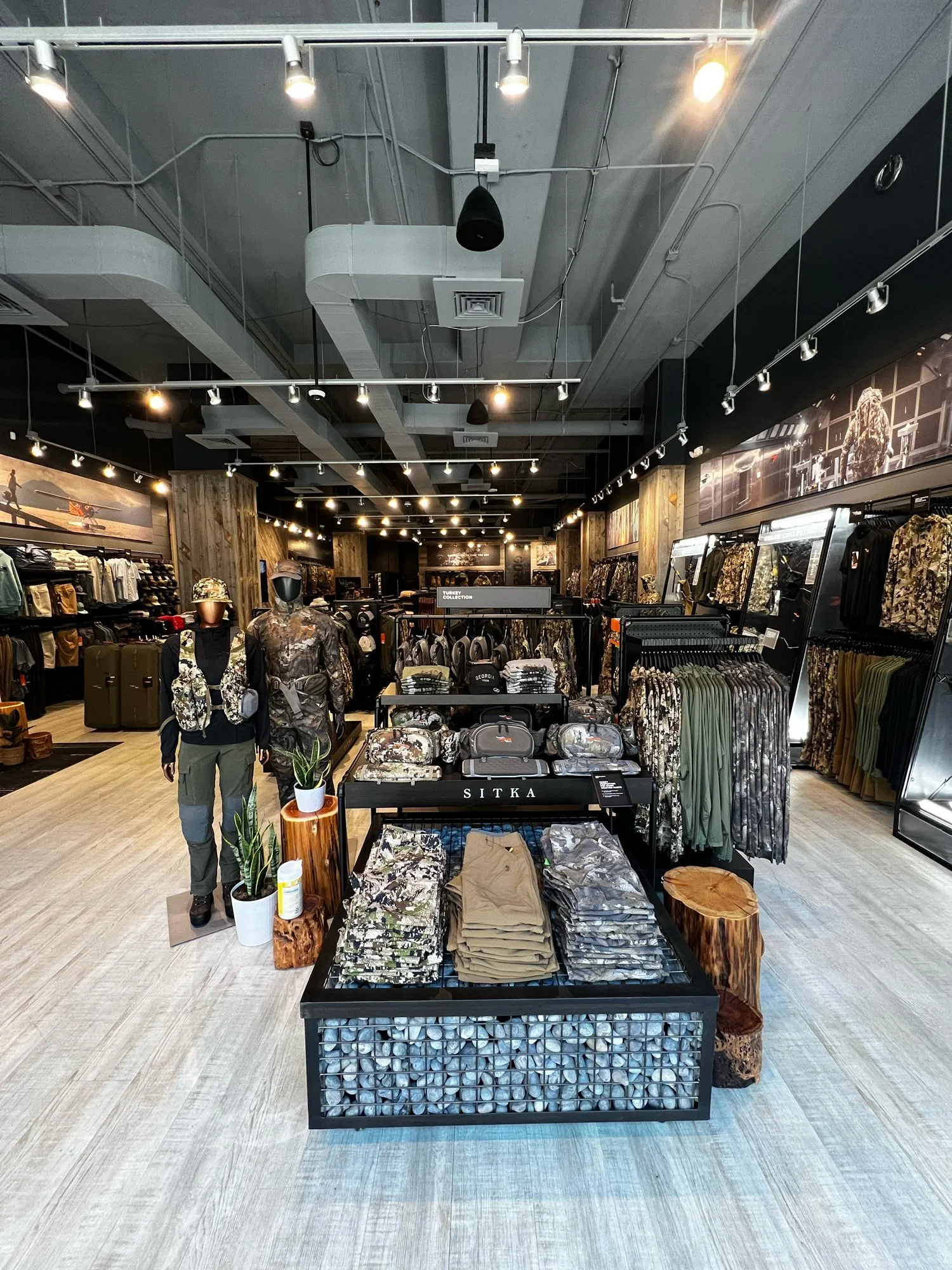 Interior of a retail store with clothing and accessories on display, including camouflage clothing, hats, and luggage, with a modern industrial ceiling and track lighting.