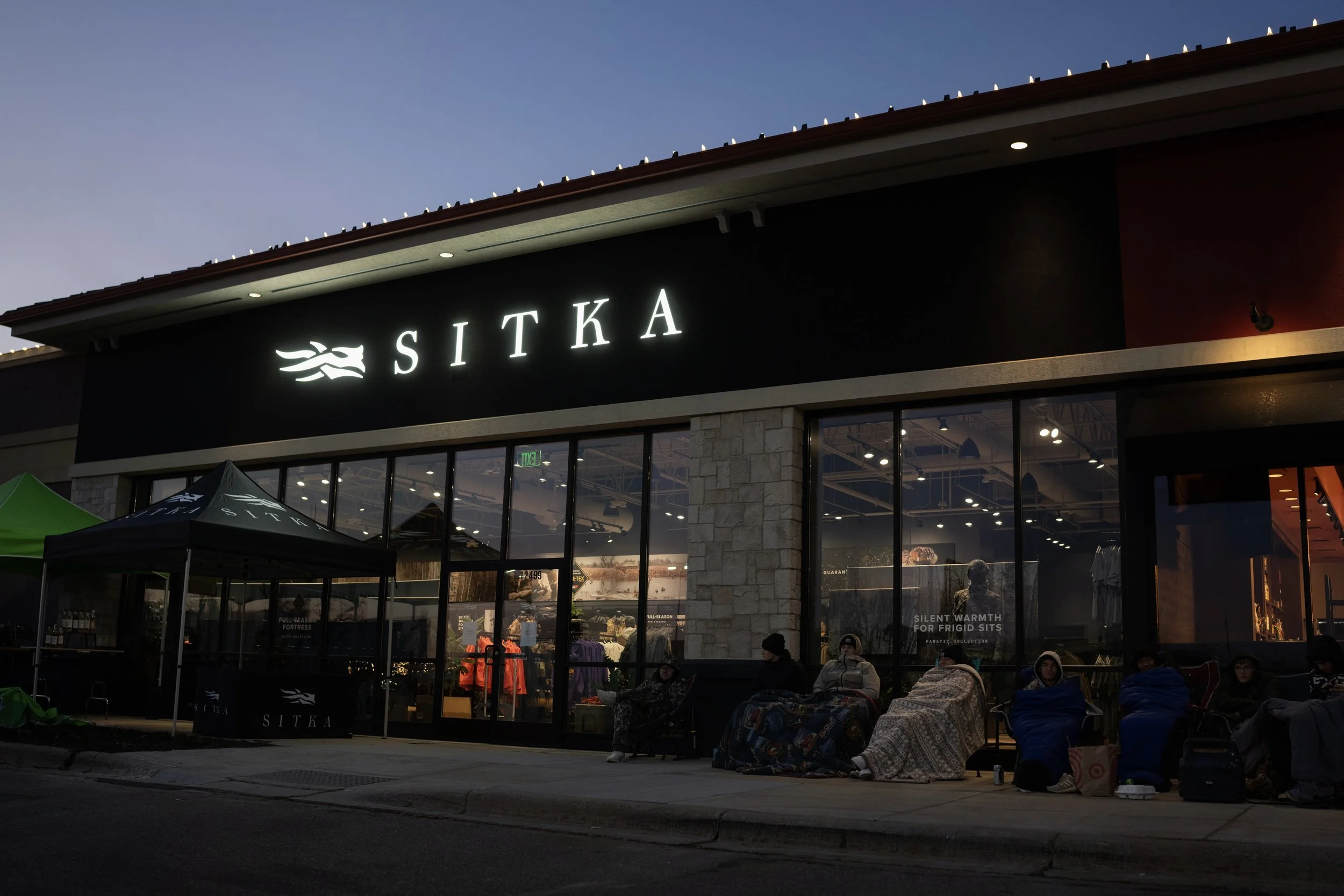 People sitting outside a retail store named SITKA during evening, some wrapped in blankets, with tents set up nearby.