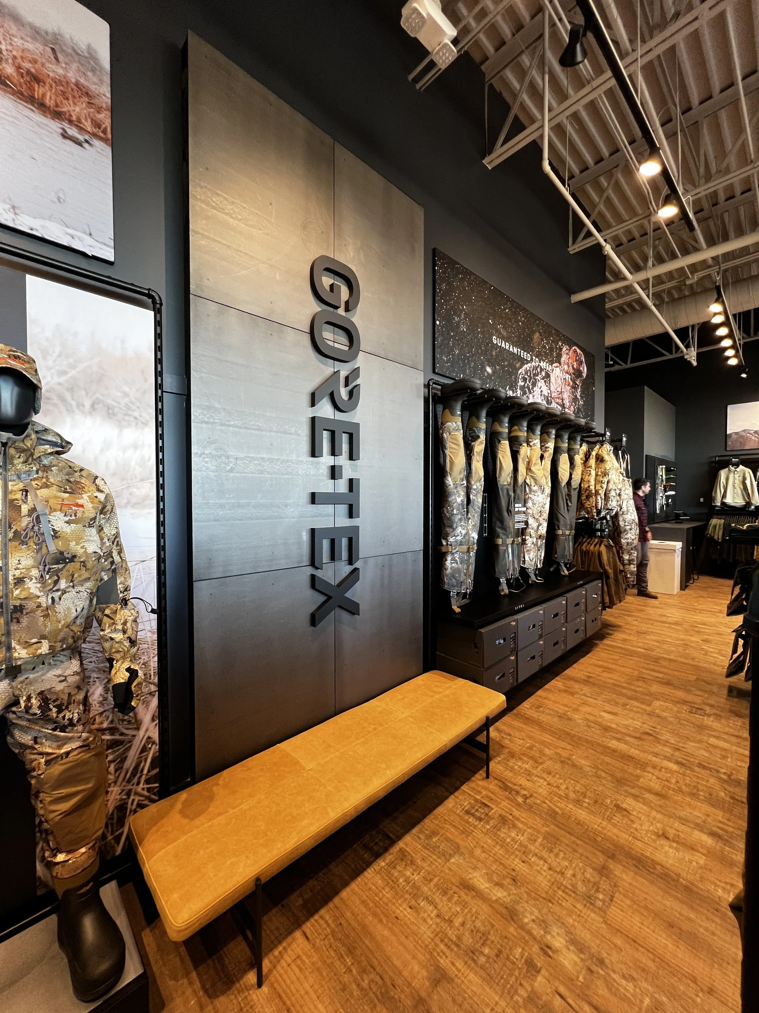 Inside a retail store displaying outdoor gear, including camouflage jackets, a bench, and advertising with the word 'OUTDOOR' on the wall.