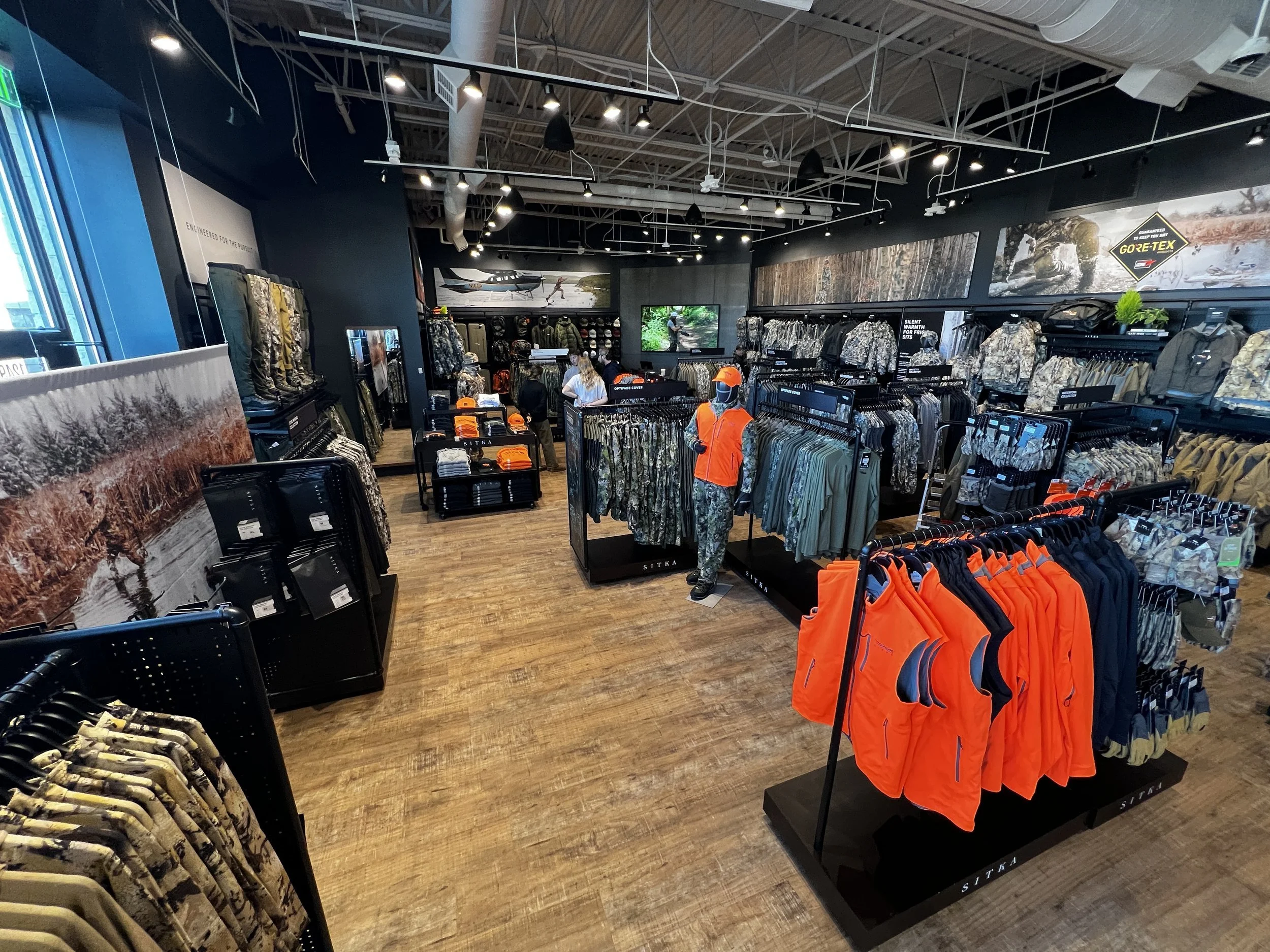Inside a retail store displaying outdoor clothing and gear, with racks of jackets, pants, and bright orange safety vests. The store has wood flooring, black walls, and television screens showing outdoor scenes and products.