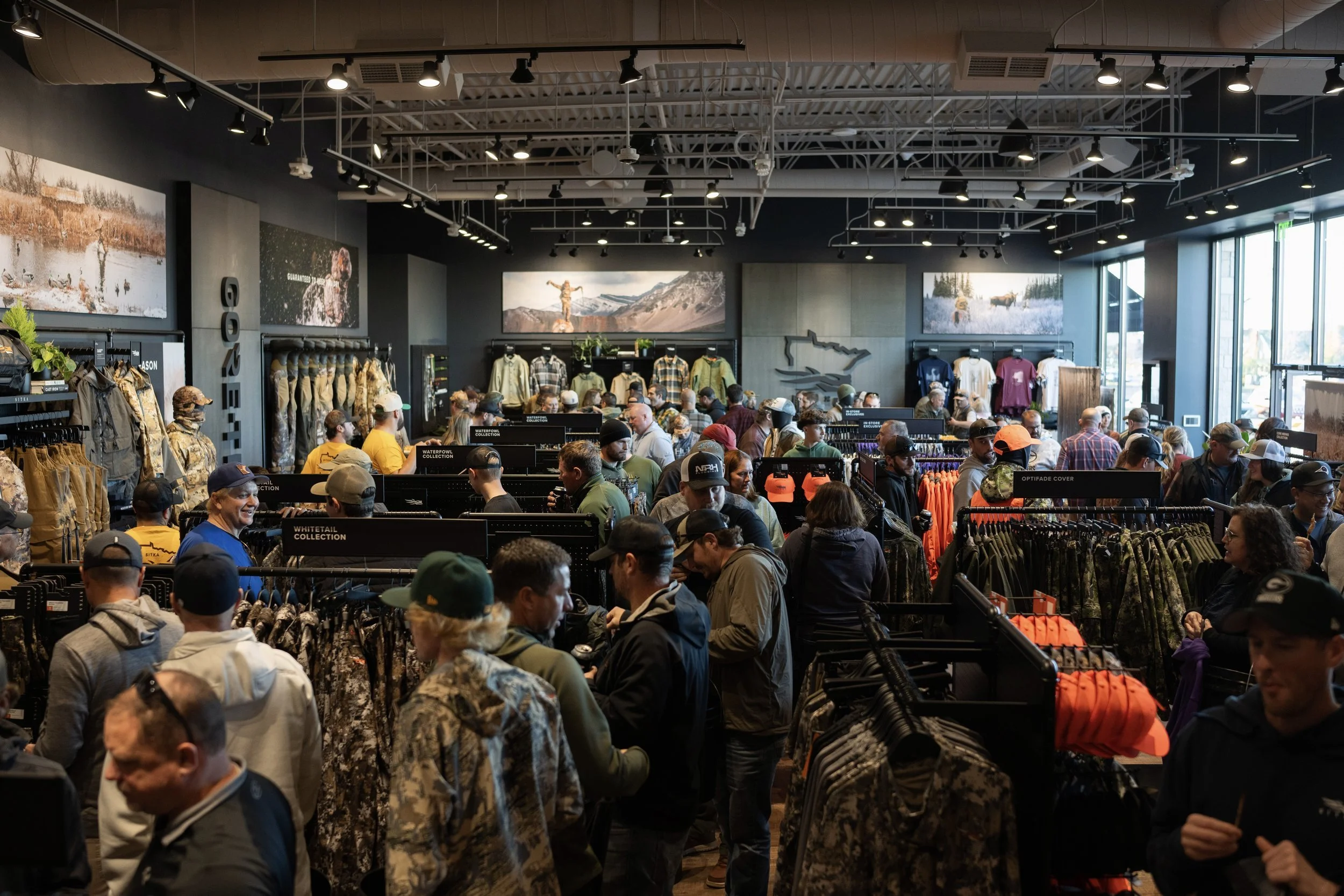 A crowded outdoor gear store filled with shoppers browsing clothing and accessories, with large windows letting in natural light and mountain landscape artwork on the walls.
