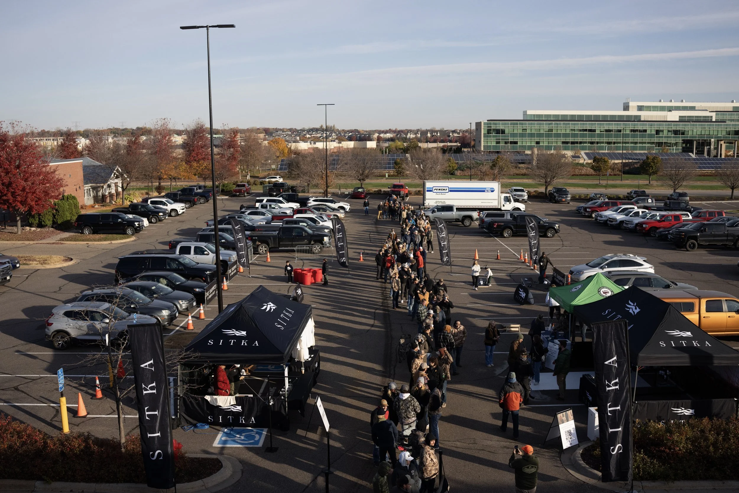 A parking lot with people walking towards tents and booths for an outdoor event on a clear day. There are trees with autumn foliage and a large building with solar panels in the background.
