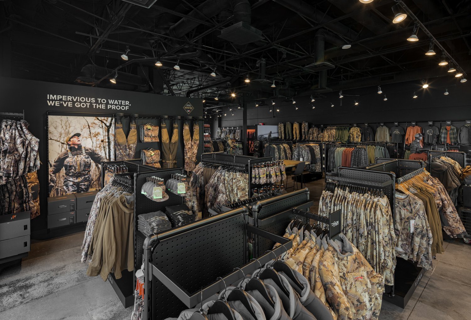 Interior of a retail store selling outdoor and military-style clothing, with racks of camouflage and rugged jackets, pants, and accessories, and a wall display featuring jackets and gear. The lighting is industrial with black ceilings and track light