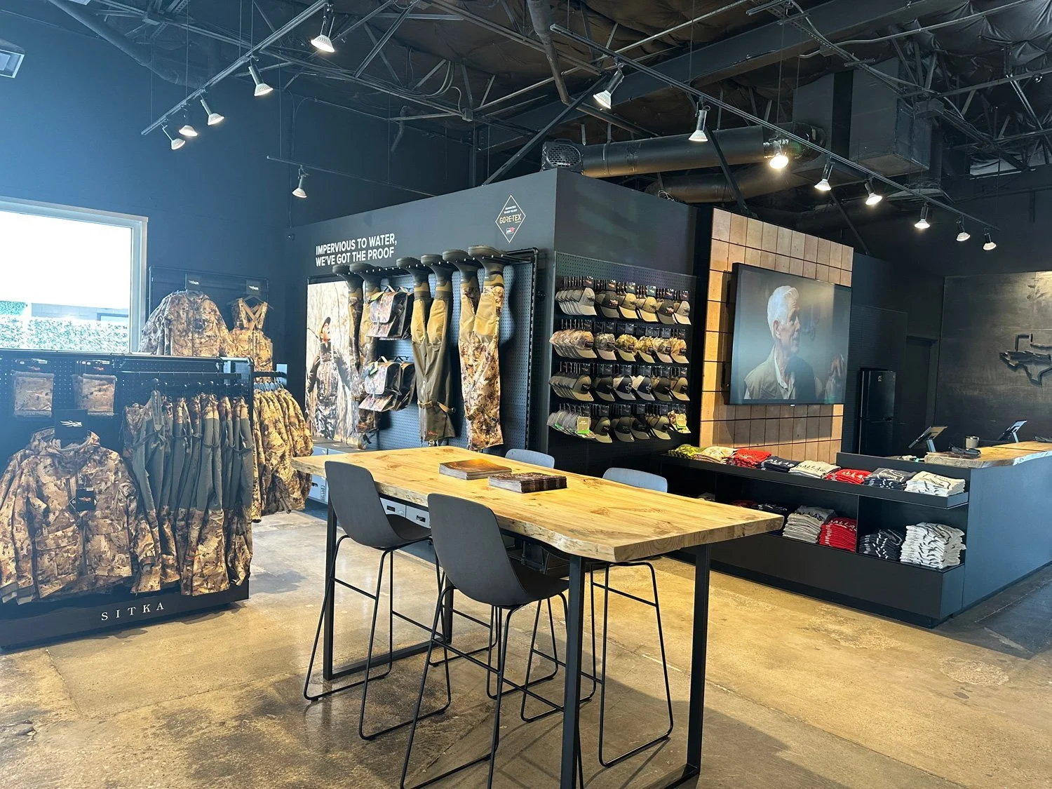 Inside a retail store with military camouflage clothing on display, a wooden table with chairs, shelves with hats and shirts, a large TV screen showing a person, and industrial lighting.