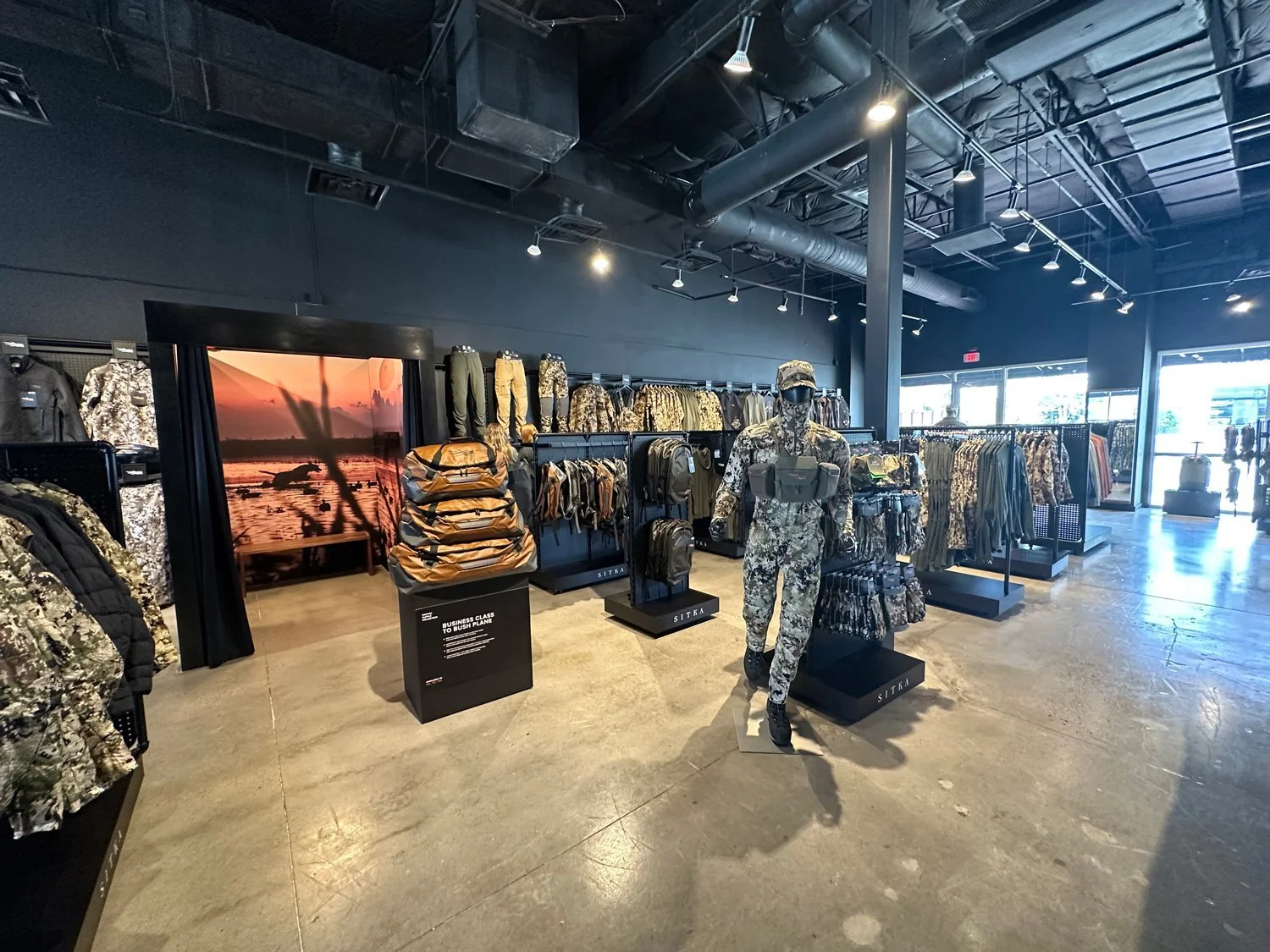 Inside a clothing store with military camouflage apparel and accessories, mannequins dressed in camouflage attire, clothing racks with camouflage jackets and pants, and a large digital display of a sunset over water.