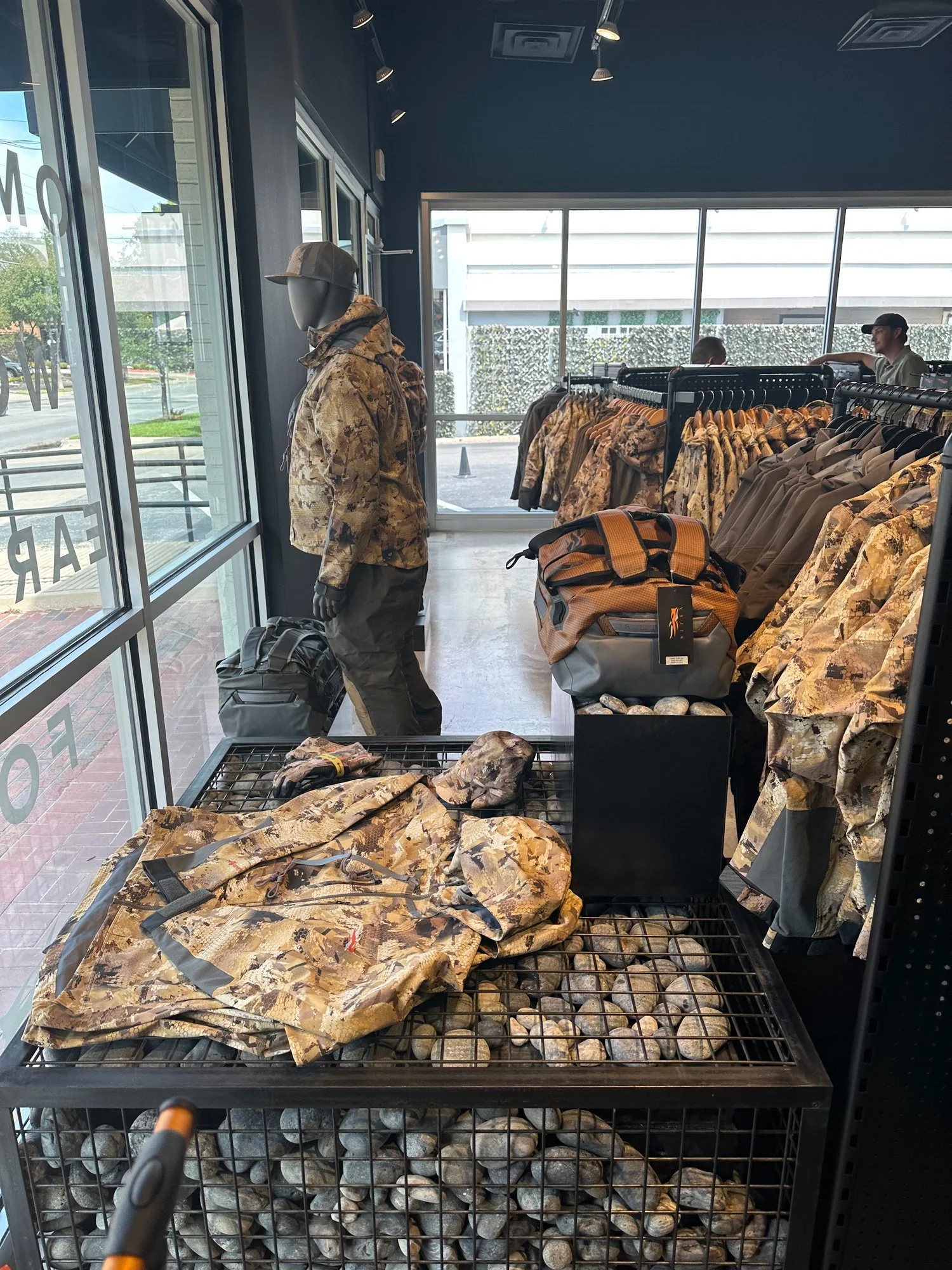 Inside a retail store with military camouflage clothing on display, including jackets and backpacks, with mannequins and shoppers visible through the large front window.