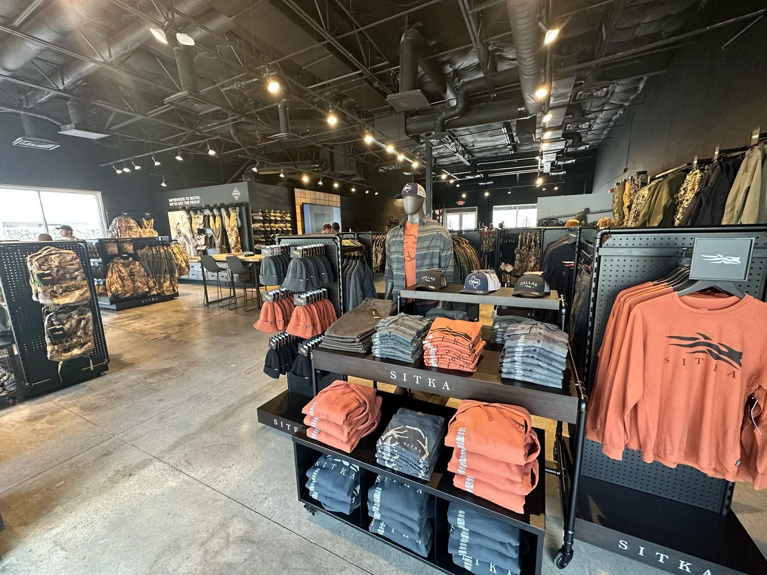 Inside a retail store with shelves and tables displaying outdoor and camouflage clothing, hats, and accessories. Black ceiling with exposed ductwork, and large windows providing natural light.