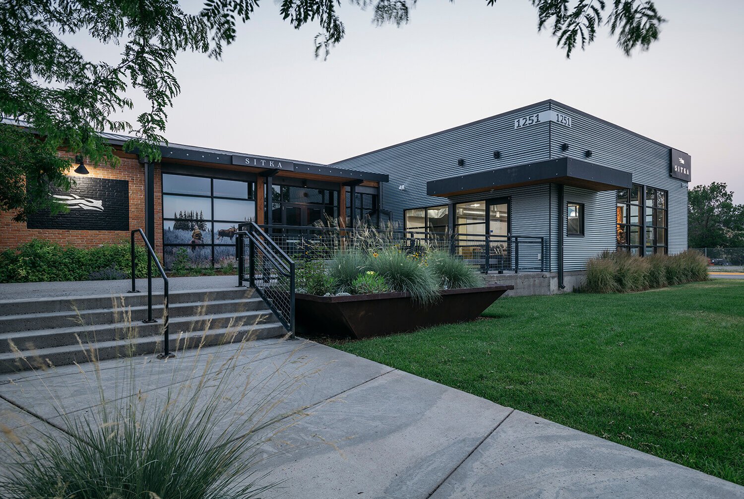 Modern commercial building with metal siding, brick accents, large glass windows, surrounding greenery, and a concrete staircase with black railings leading to the entrance.