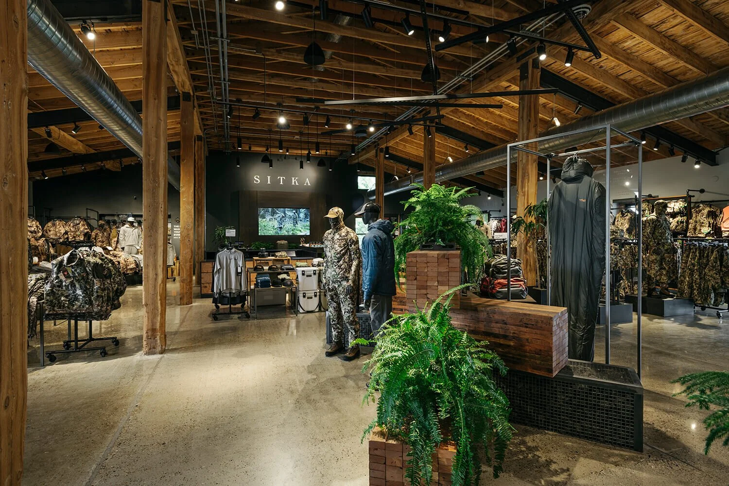 Interior of a clothing store with wooden beams, mannequins dressed in camouflage and outdoor apparel, and green plants.