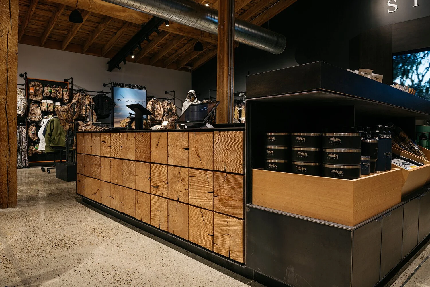 Interior of a retail store with a rustic wooden checkout counter and outdoor gear on display, including backpacks, jackets, and water bottles.