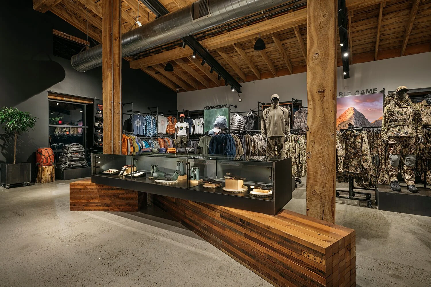 Interior of a clothing store with outdoor and athletic wear, mannequins dressed in outdoor gear, wooden accents, exposed ceiling beams, and display shelves with clothing, accessories, and backpacks.
