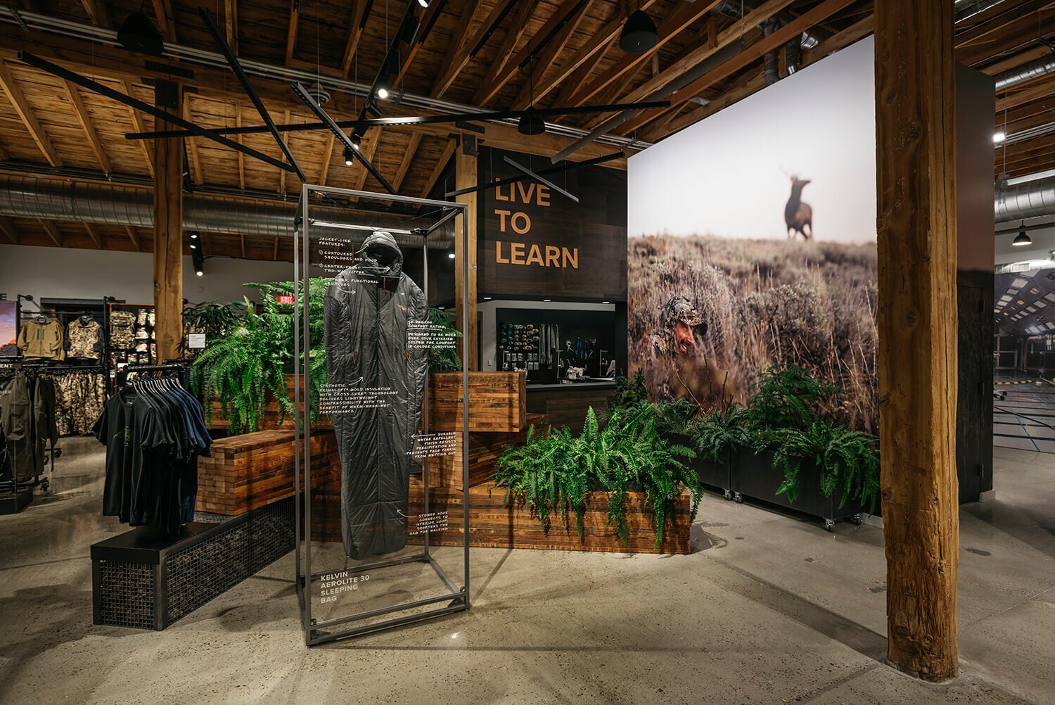 Outdoor clothing store display featuring a black sleeping bag in a glass case, surrounded by green plants and a large framed photo of someone in camouflage with a dog in a field, and the phrase 'LIVE TO LEARN' on the wall.