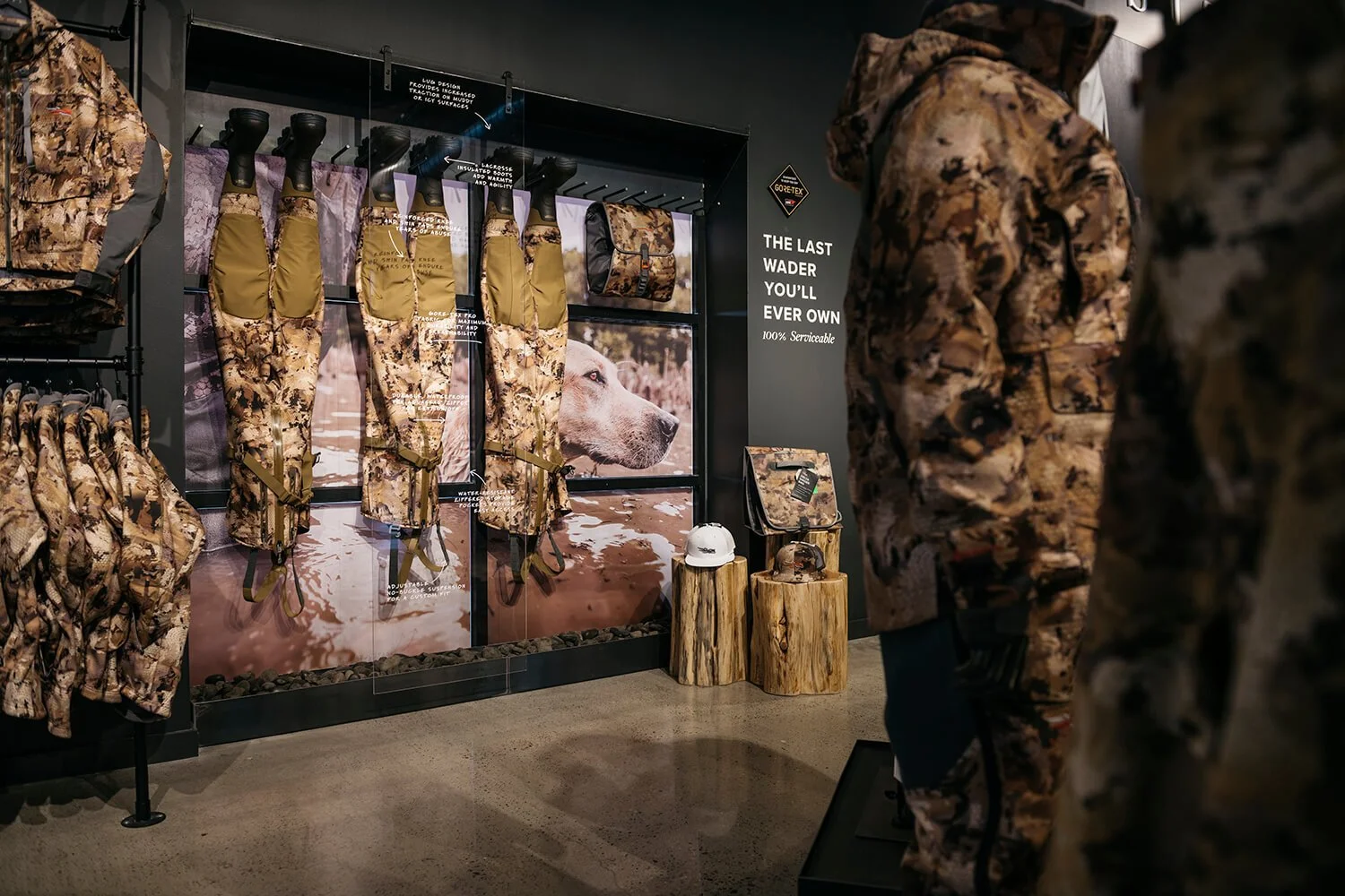 Display of outdoor hunting gear with camouflage jackets, bibs, and accessories in a store. A wall features a large image of a hunting dog and signs indicating product features. Manikins dressed in camouflage and outdoor gear are visible, along with n