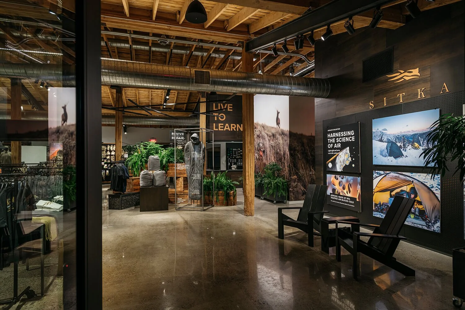 An indoor retail space with wooden beams and a polished concrete floor, featuring wall art, plants, mannequins, and displays related to outdoor activities and gear.