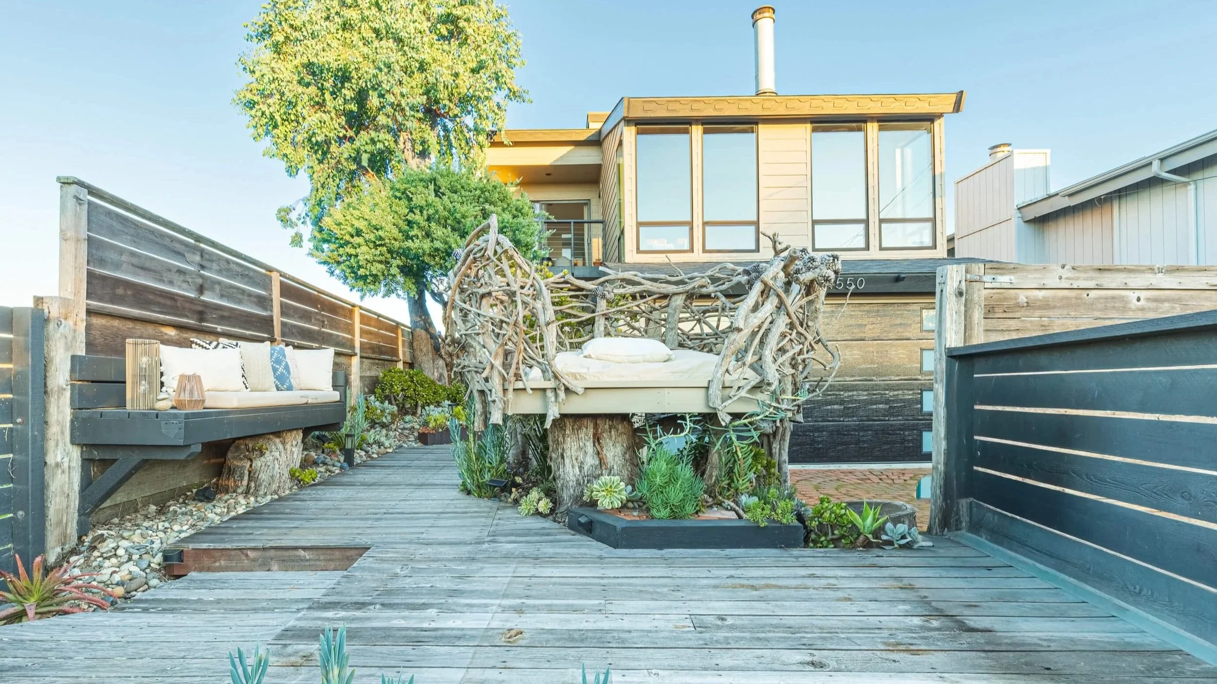An outdoor patio area featuring a rustic sofa with pillows, a unique bed-like seating surrounded by driftwood, and a variety of succulents and plants, with a modern house and wooden fencing in the background.