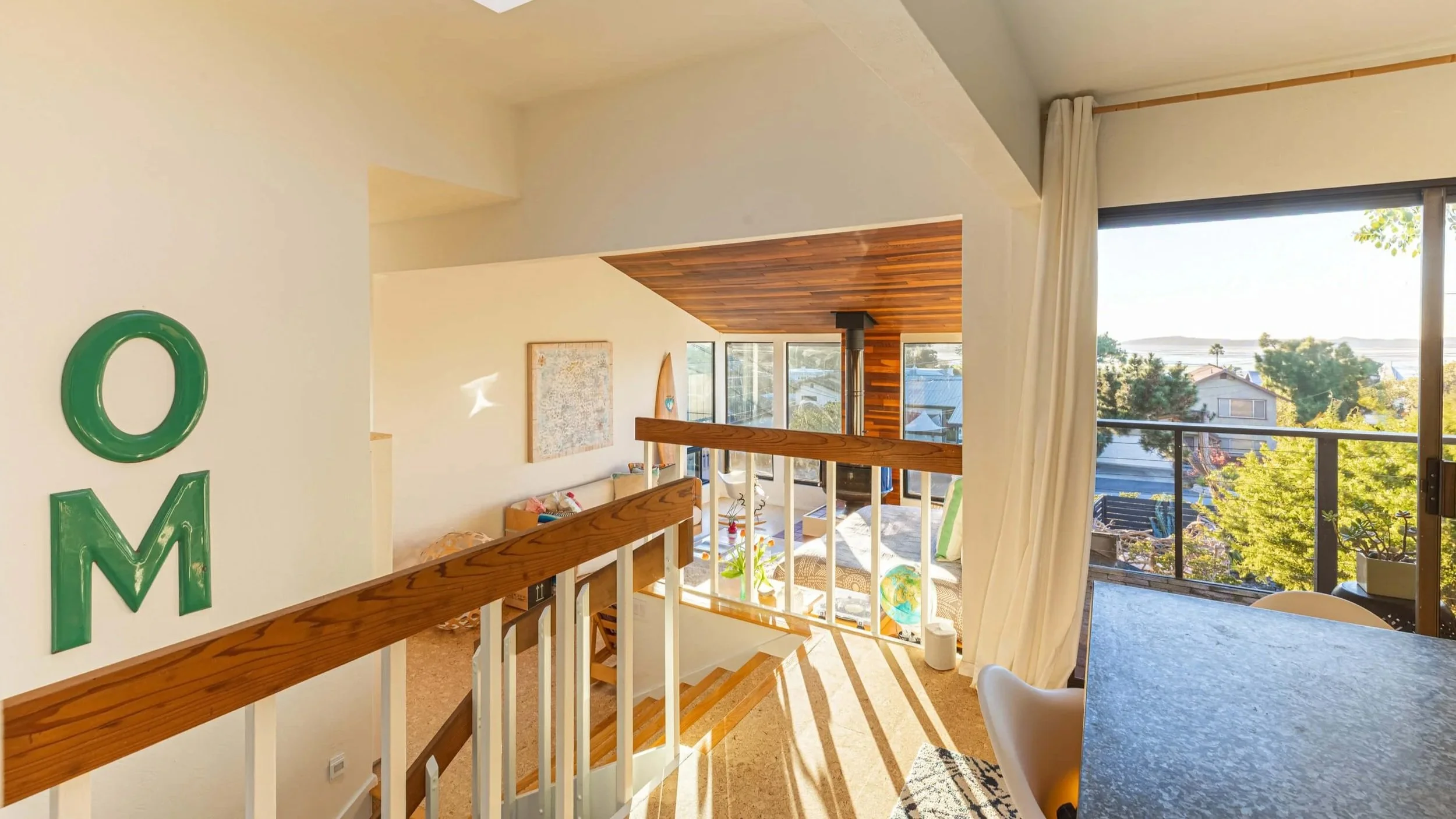 Bright living space with large windows showing trees and houses outside, wooden staircase, artwork on wall, and seating area with a globe and cushions.
