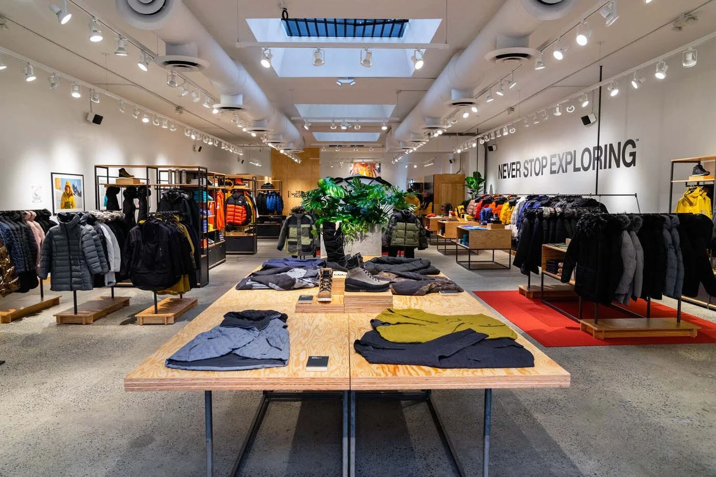 Clothing store interior with jackets and outdoor apparel on racks and tables, including a sign with 'NEVER STOP EXPLORING' on the wall.
