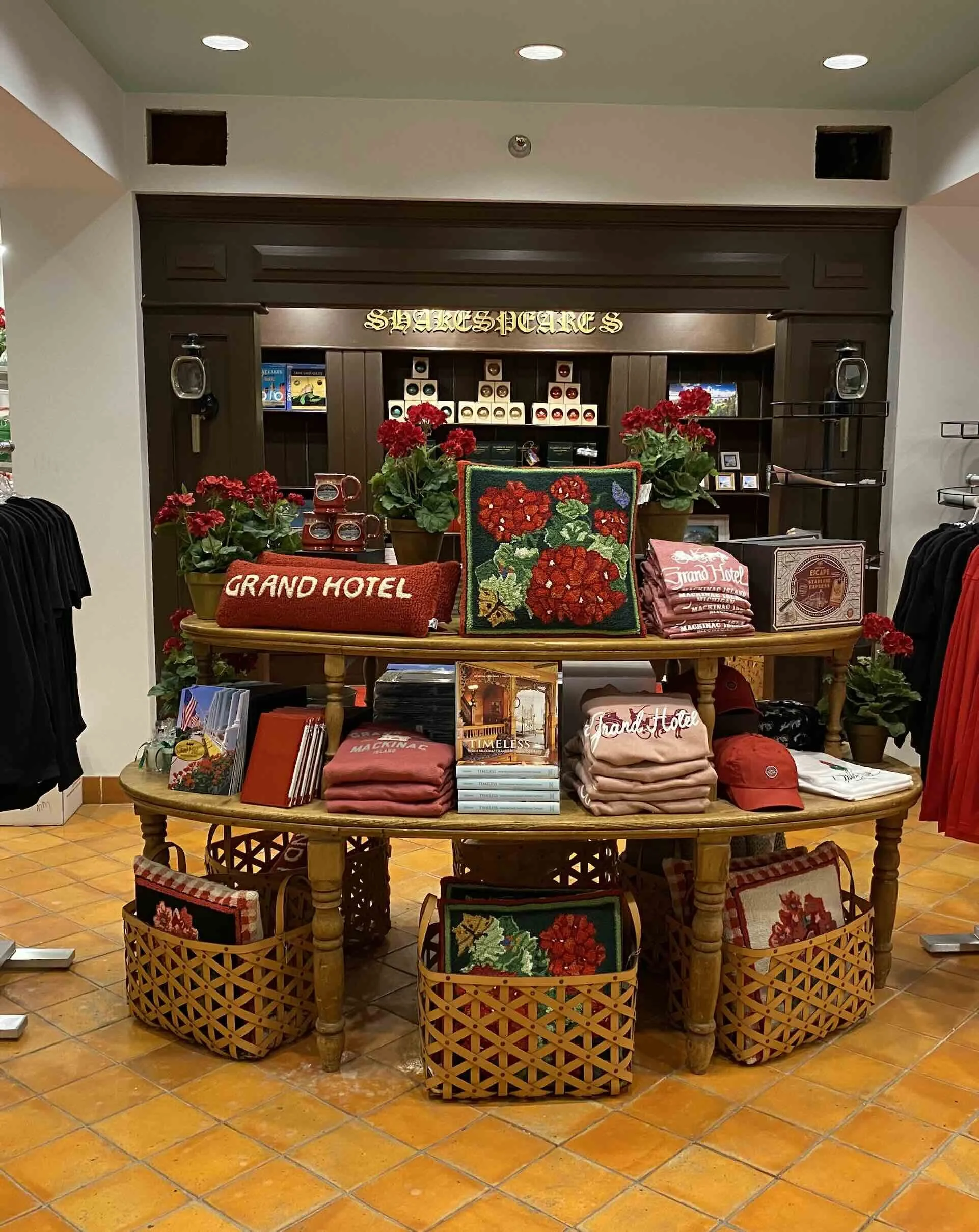 Display table with red flowers, embroidered pillow, and folded clothing in a store with a sign reading 'Shakespeare' in the background.