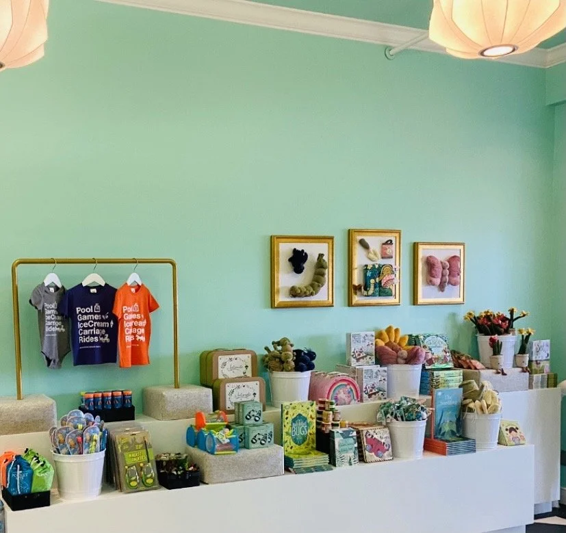 Display of children's toys, books, and clothing in a brightly decorated store with framed art on the wall.