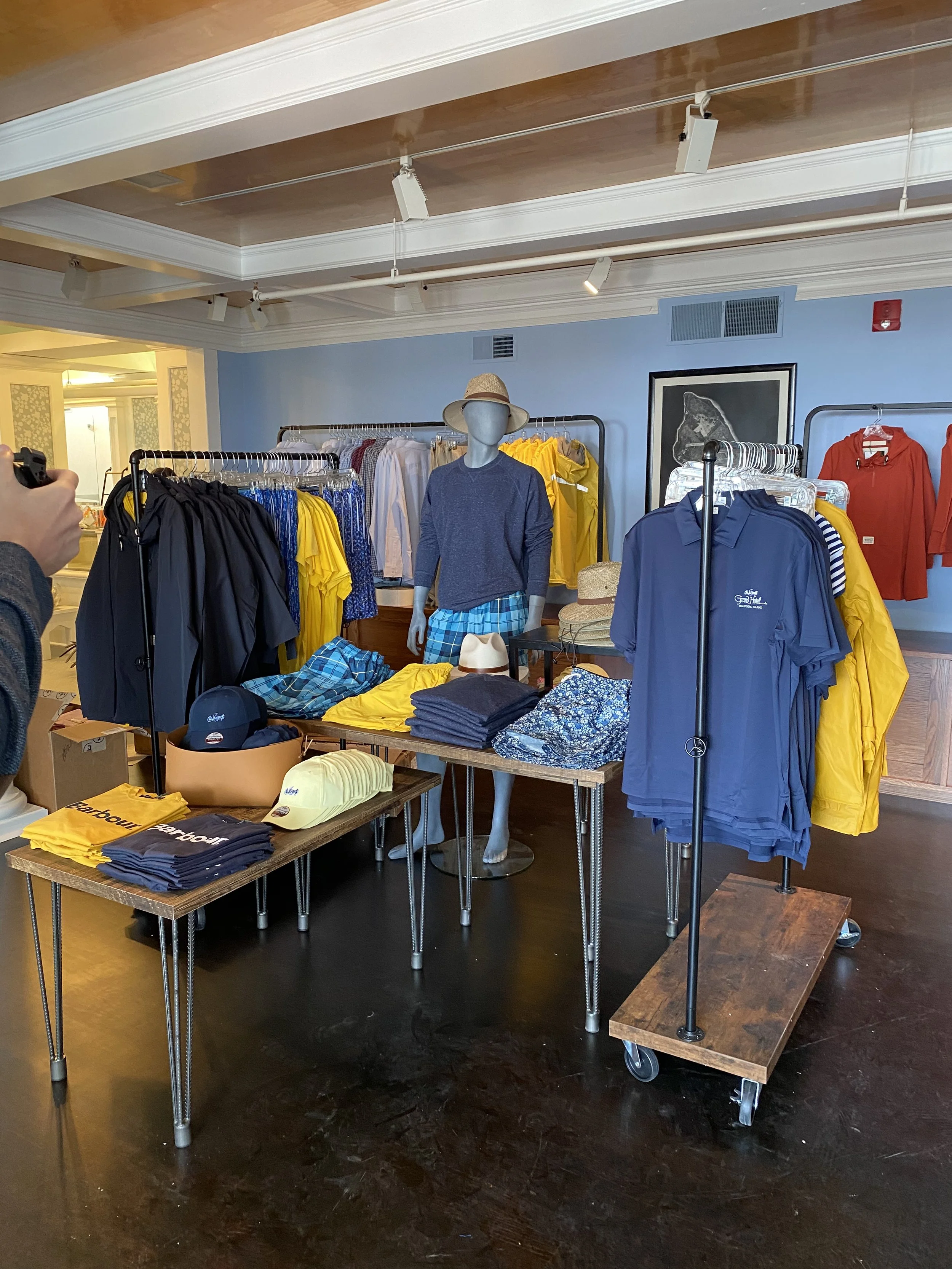 Clothing store display with mannequins wearing casual clothes, racks of various jackets and shirts, and accessories like hats on tables.