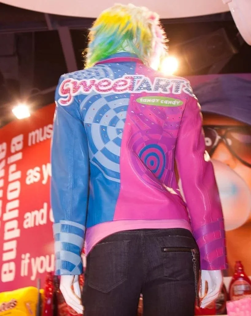 Back of a mannequin with rainbow-colored hair wearing a multicolored jacket featuring pink, blue, and purple sections with various designs, including the words 'sweetART' and 'tangy candy.' The mannequin is in a store with red and white signage in the background and dark ceiling with lights.