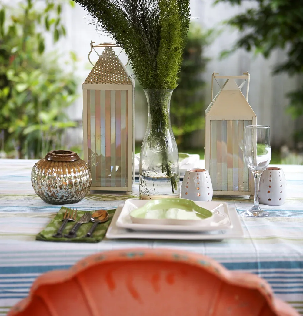 A decorated outdoor dining table with a striped tablecloth, centerpiece of green plants in a glass vase, white lanterns, a silver vase, a glass of water, and white candle holders with orange accents.