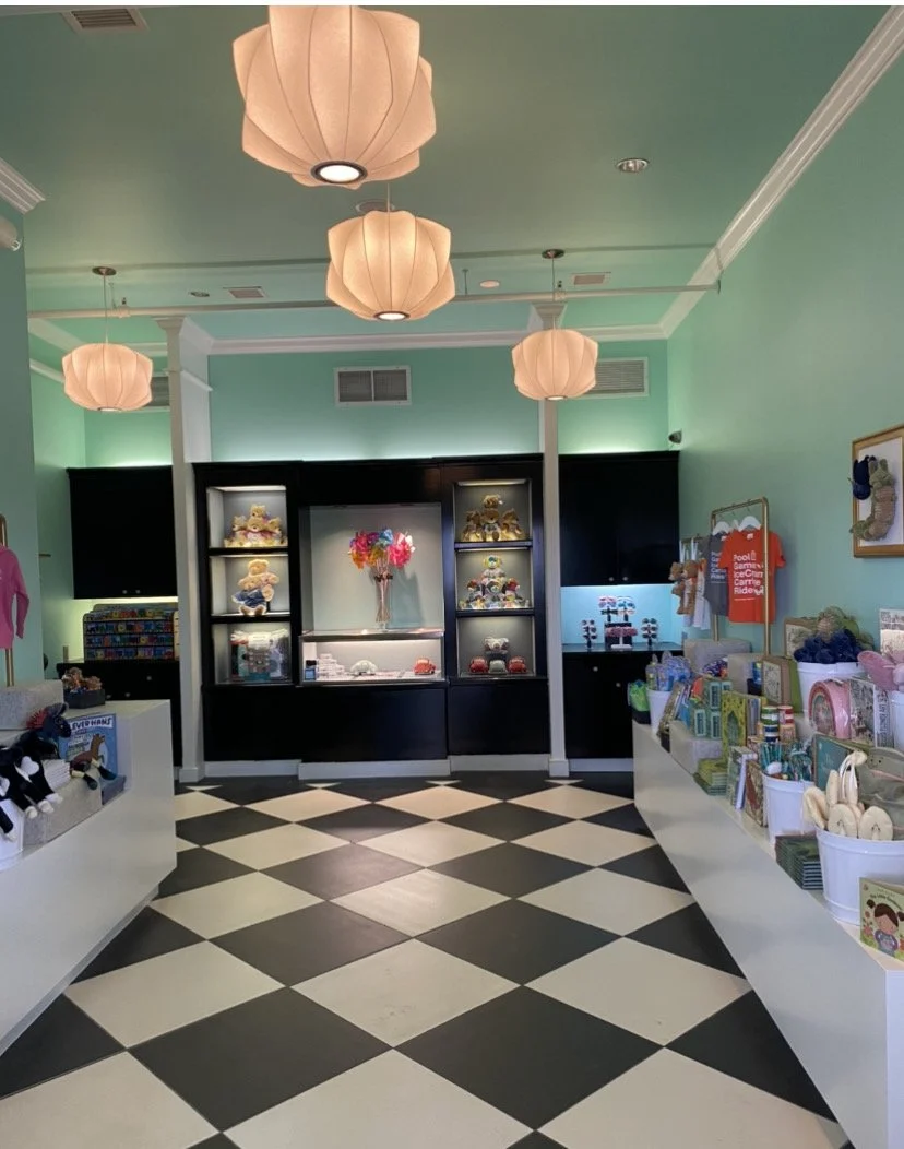 Gift shop interior with black and white checkered floor, mint green walls, and pink paper lantern ceiling lights; display shelves with toys, plush animals, and greeting cards.
