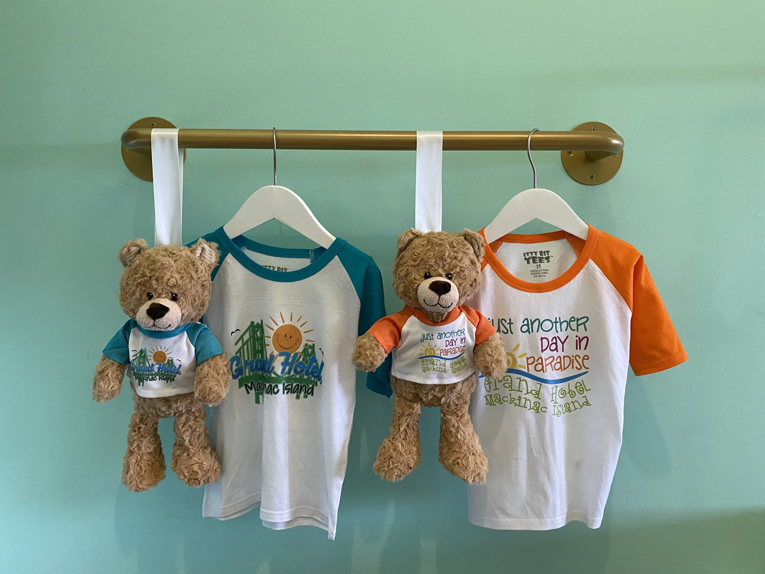 Three teddy bears hanging on white and orange T-shirts on a hanger against a mint green wall.