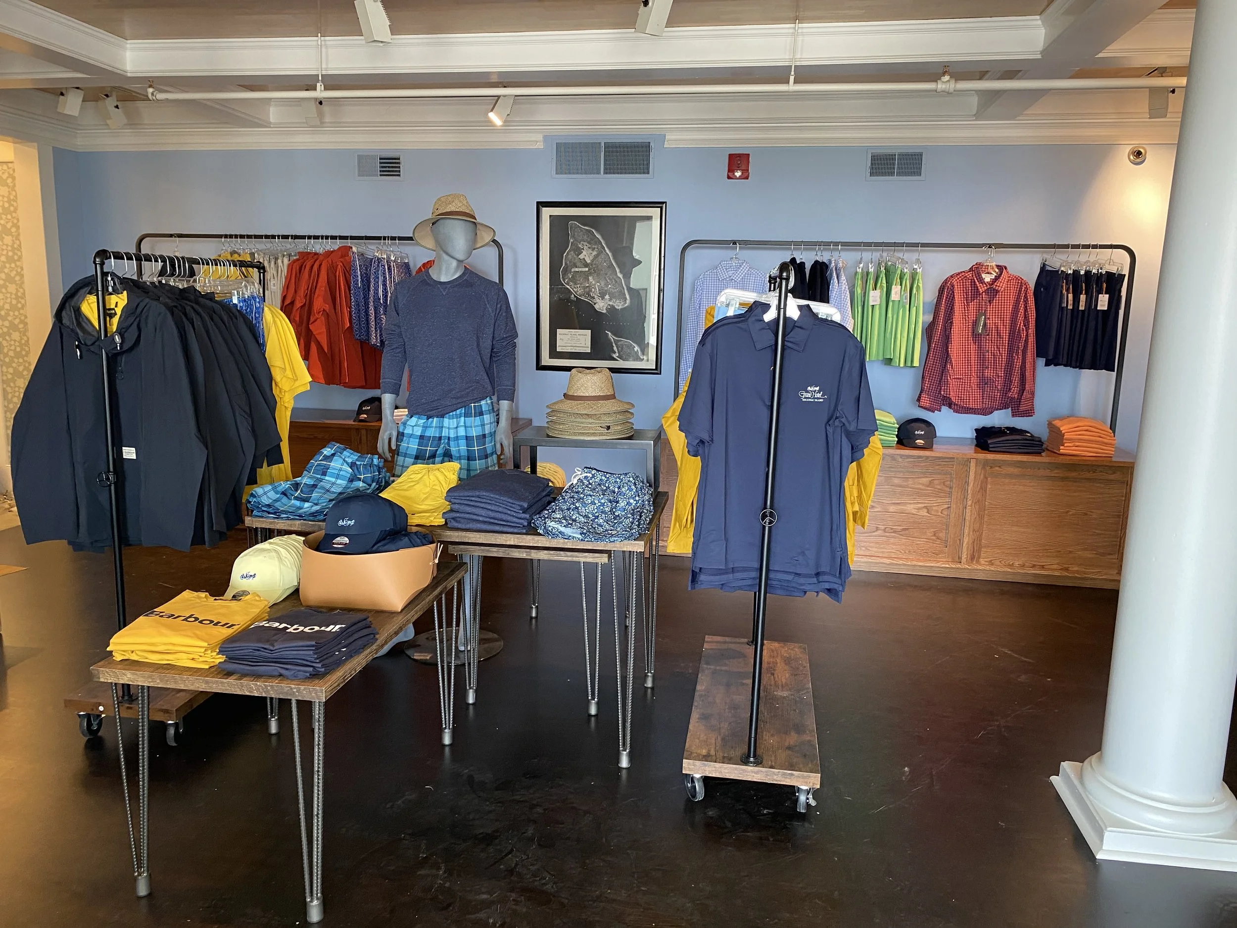 Clothing display in a retail store with racks of colorful shirts and jackets; mannequins dressed in casual attire; a framed map on the wall; tables with folded clothes and accessories; bright lighting and polished wooden floors.