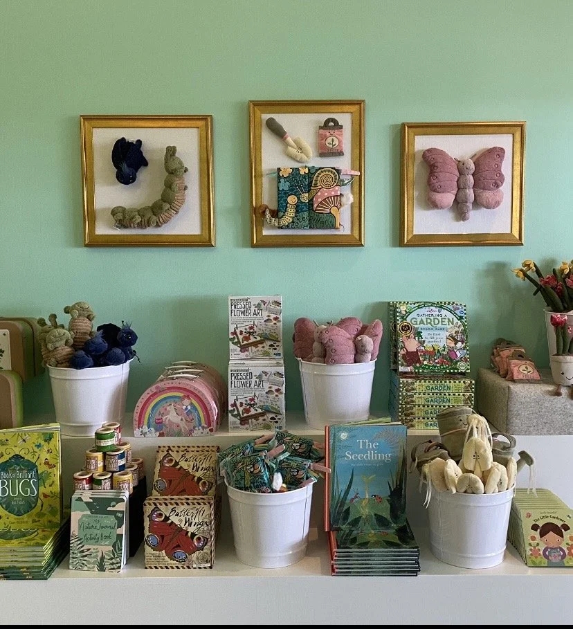 Display of children's books and toys, including stuffed animals, on a table in front of green wall with framed art pieces.