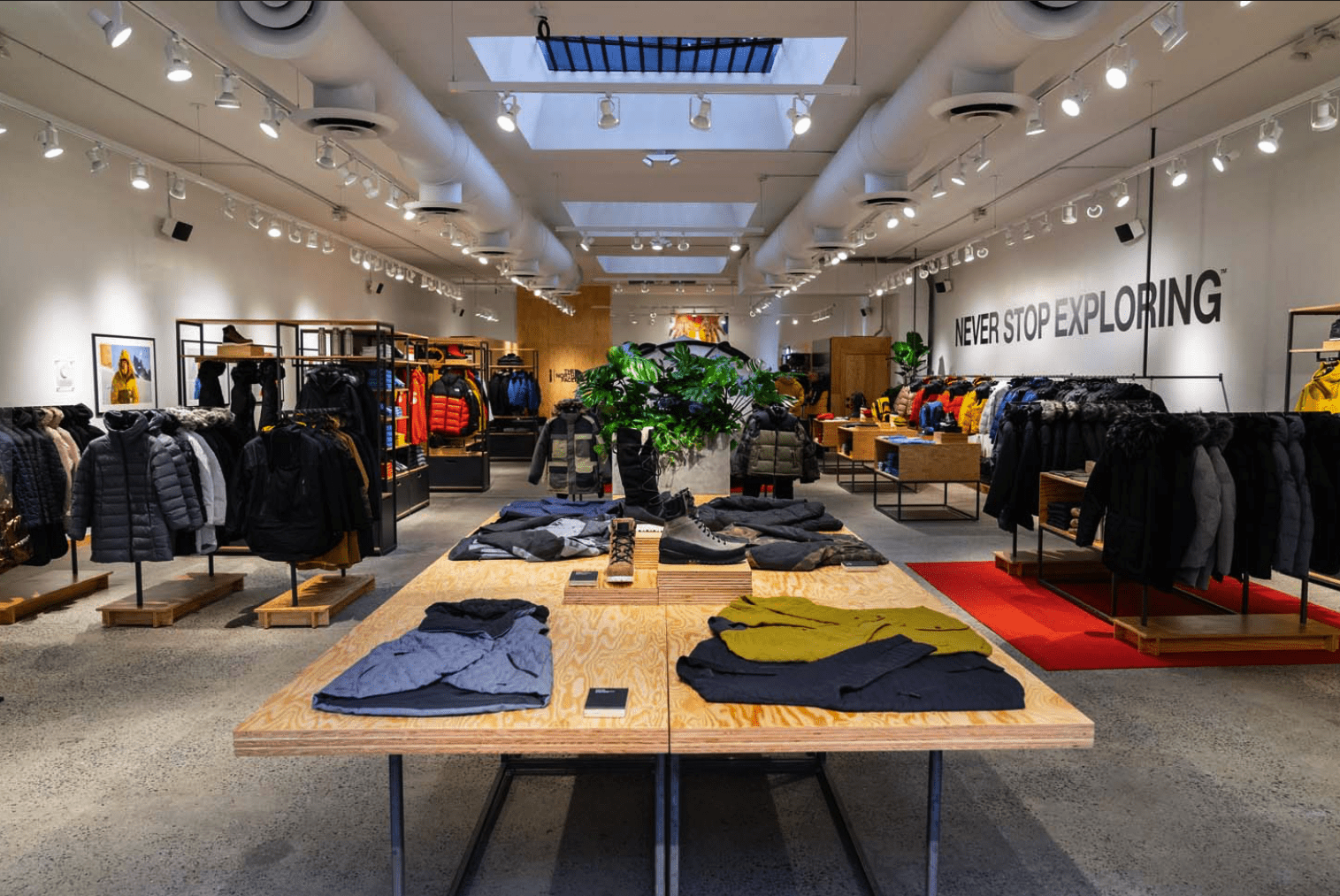 A clothing store displaying jackets, sweaters, and outdoor gear on racks and tables, with the phrase "Never Stop Exploring" on the wall.