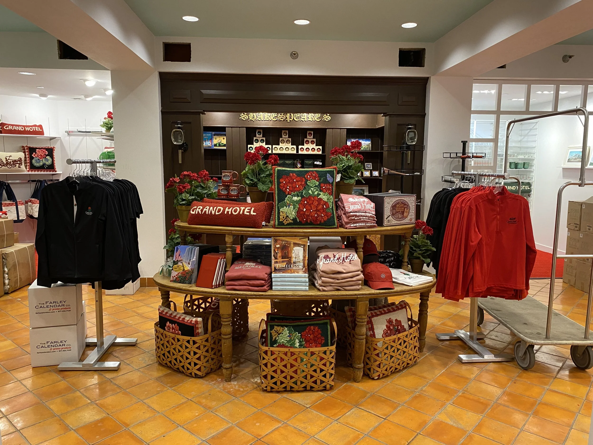 A retail store display featuring decorative items and souvenirs related to the Grand Hotel, including pillows, towels, and artwork, with shelves and racks of clothing and other merchandise in the background.