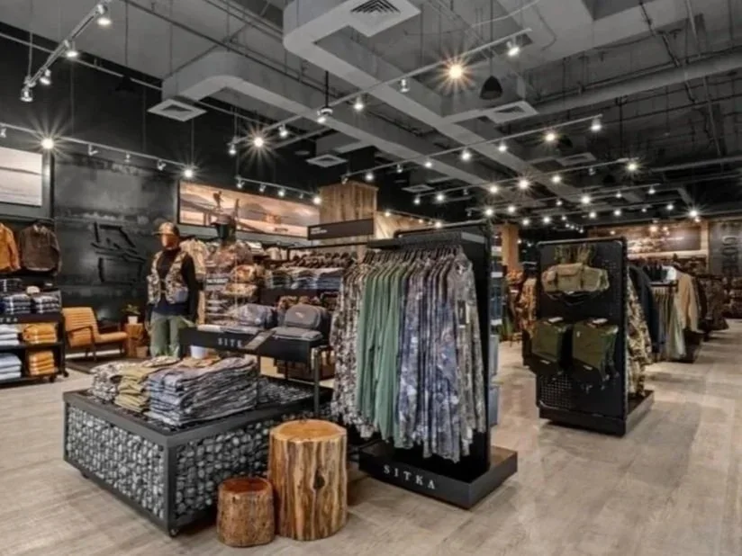 A retail clothing store with racks of folded and hanging clothes, including shirts and jackets, illuminated by ceiling spotlights, and a seating area with chairs and wooden stools.
