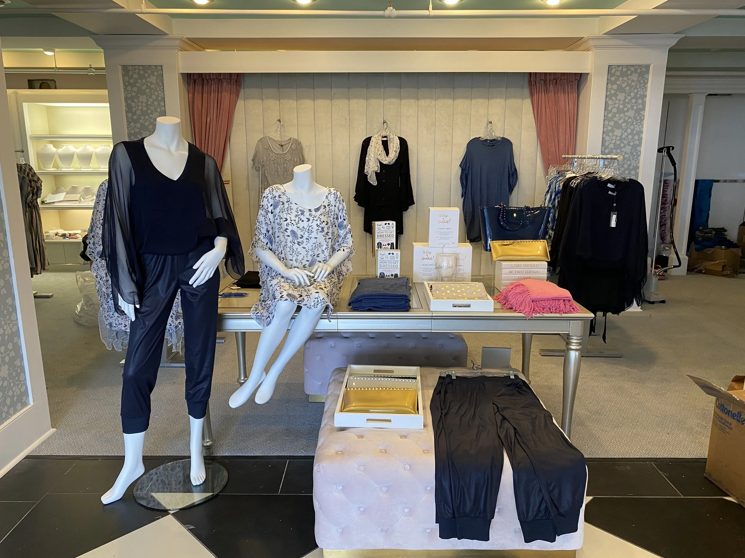 A retail display features mannequins dressed in black and floral clothing, with accessories like bags and scarves, in a boutique setting with shelves of jewelry in the background.