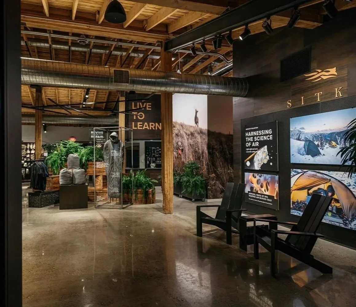 Interior of an adventure or outdoor gear store with wooden beams, potted plants, display mannequins, and large posters about air science and outdoor activities, with black chairs in the foreground.