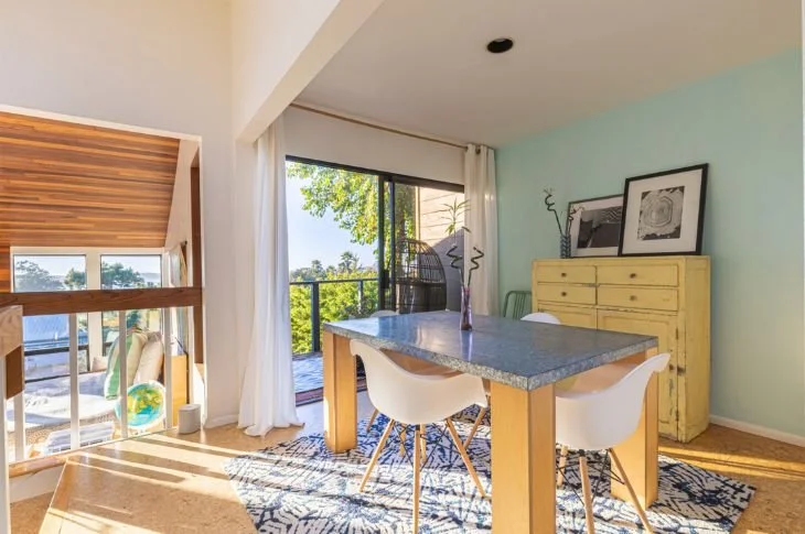 Bright living room with large window and sliding glass door leading to a balcony, wooden and glass railing, blue accent wall, yellow dresser with framed artwork, white chairs around a table with a gray tabletop, wicker chair on balcony, modern indoor
