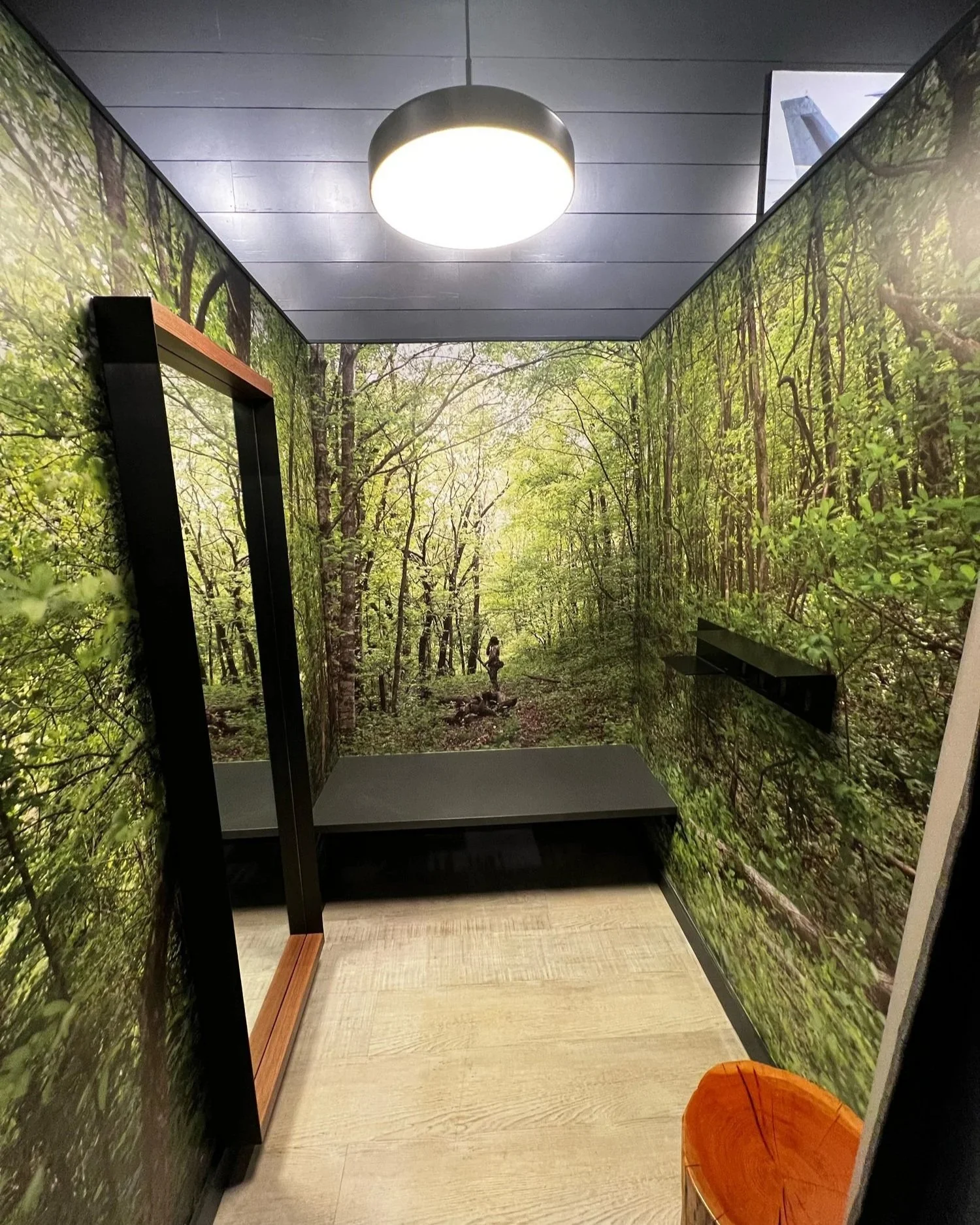 A small room with forest-themed wallpaper depicting a lush green forest, with a round ceiling light illuminating the space, and a simple wooden bench and an orange chair in the corner.