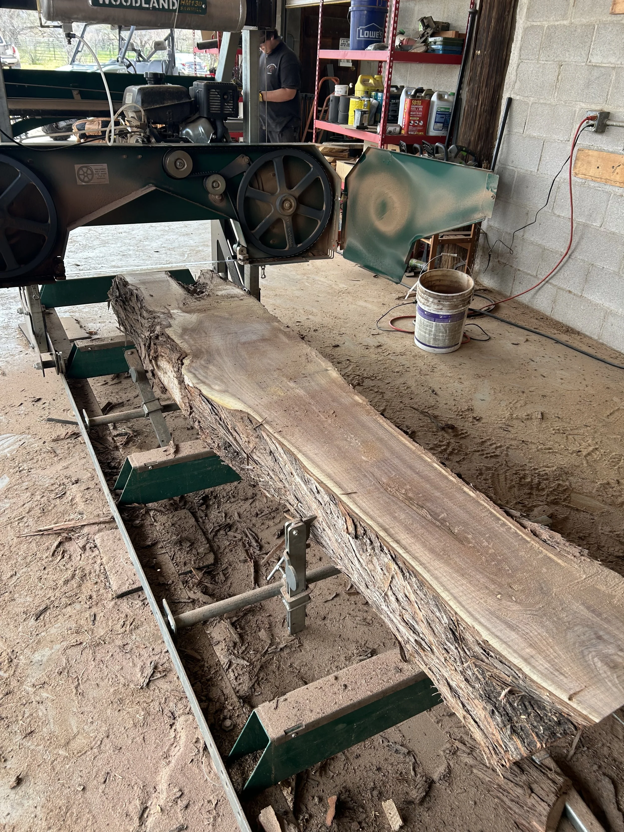 A woodworking workshop with a large piece of rough-cut wood on a saw guide, with a sawmill blade and mechanisms visible. In the background, a person is working near shelves holding various containers and tools.