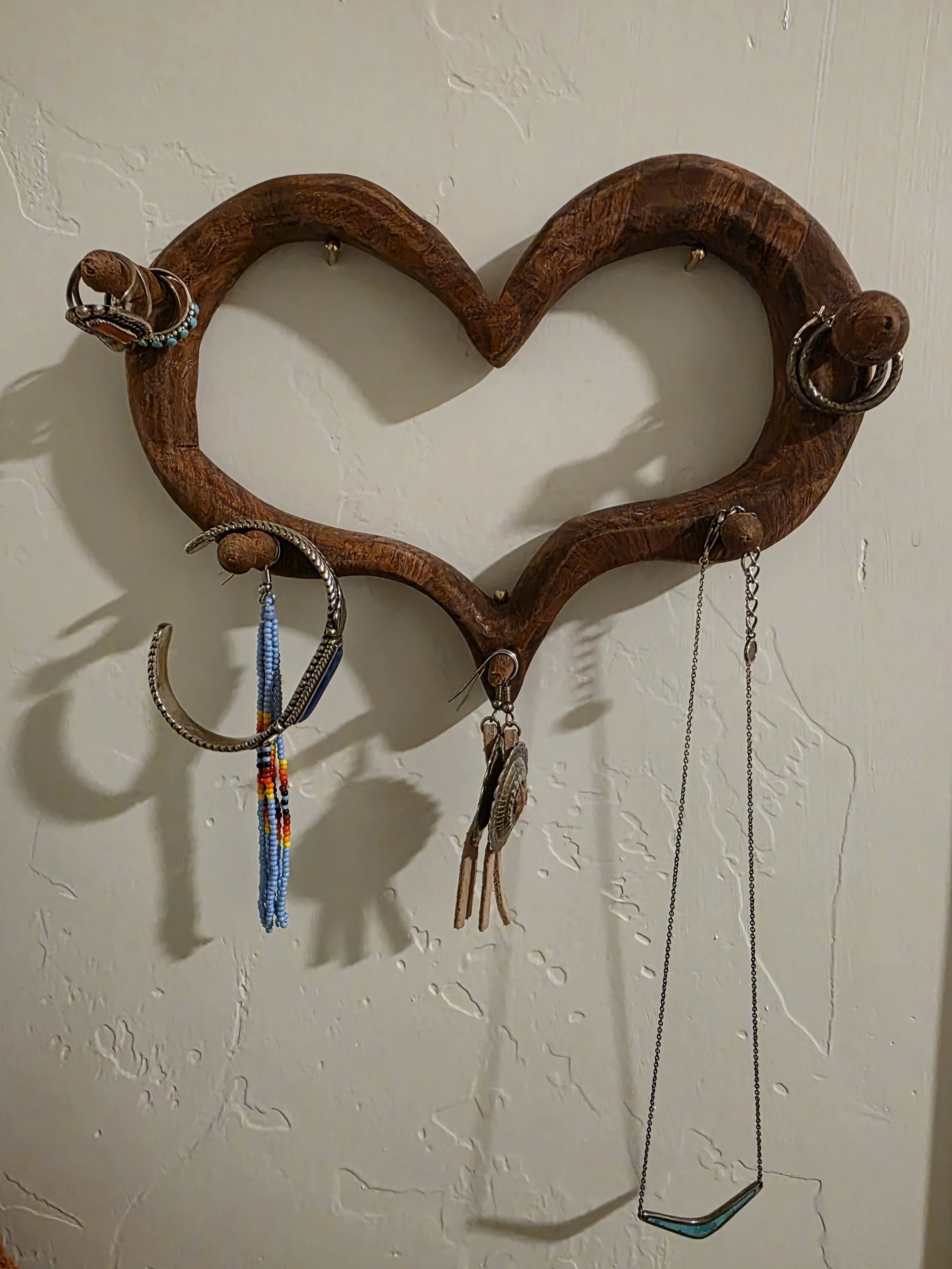 Wooden jewelry holder shaped like a heart attached to a textured, off-white wall. It holds various earrings, necklaces, and a bracelet.