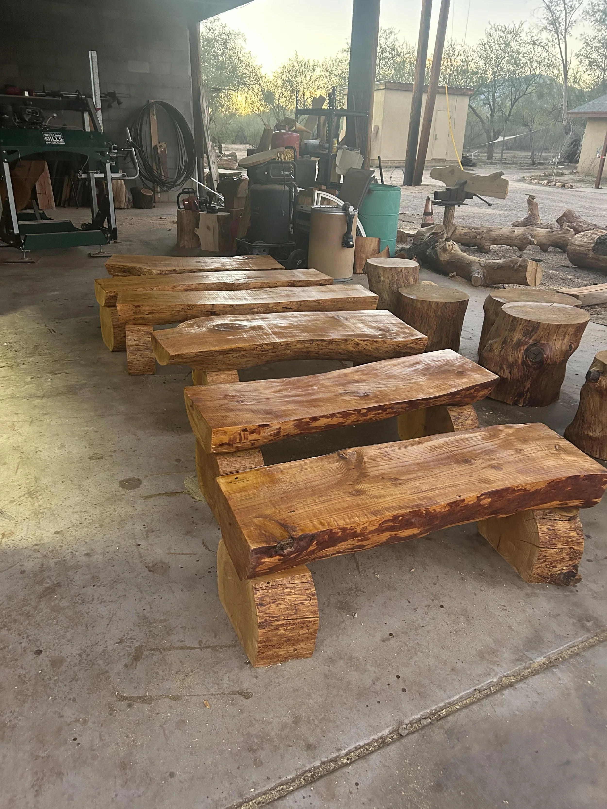 Stacked wooden benches made from large, natural wood logs with a rustic appearance in a workshop with outdoor scenery visible through the open entrance.