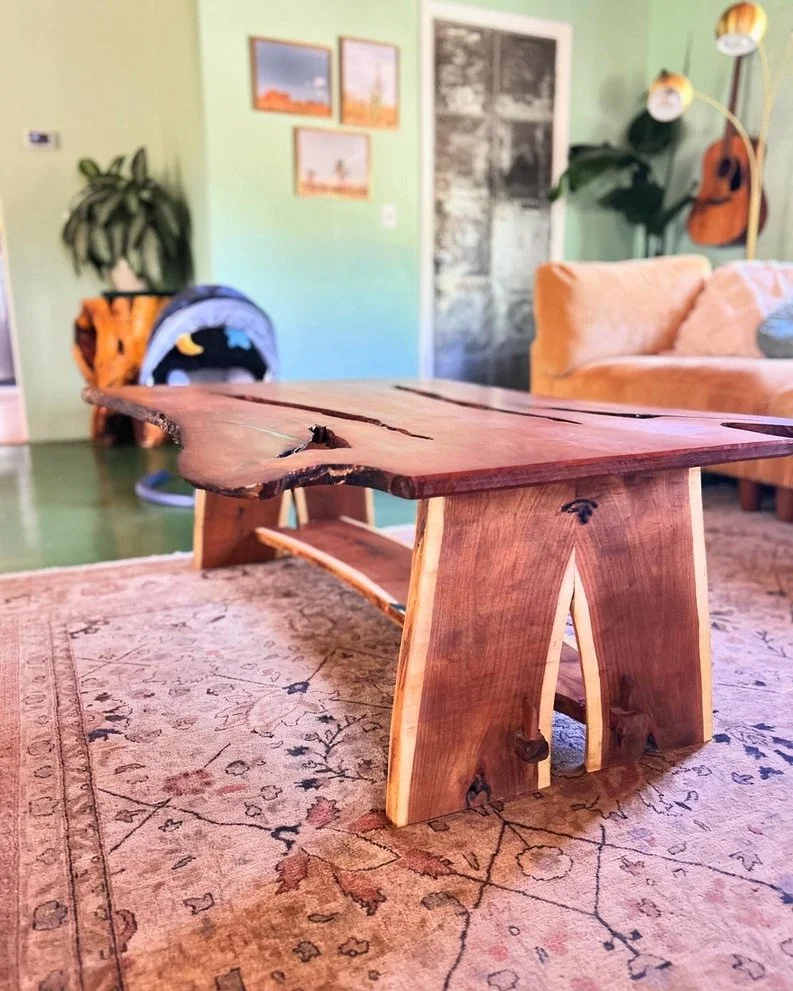 A rustic wooden coffee table on a patterned area rug in a cozy living room, with a green wall, framed pictures, a beige couch, a guitar, and a houseplant in the background.