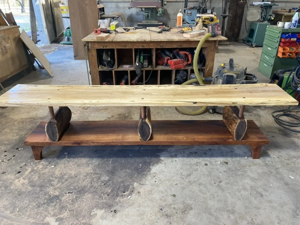 A woodworking workshop with a large, partially assembled wooden table, featuring a lighter wood top and a darker wood base with arched legs, surrounded by various tools and equipment.